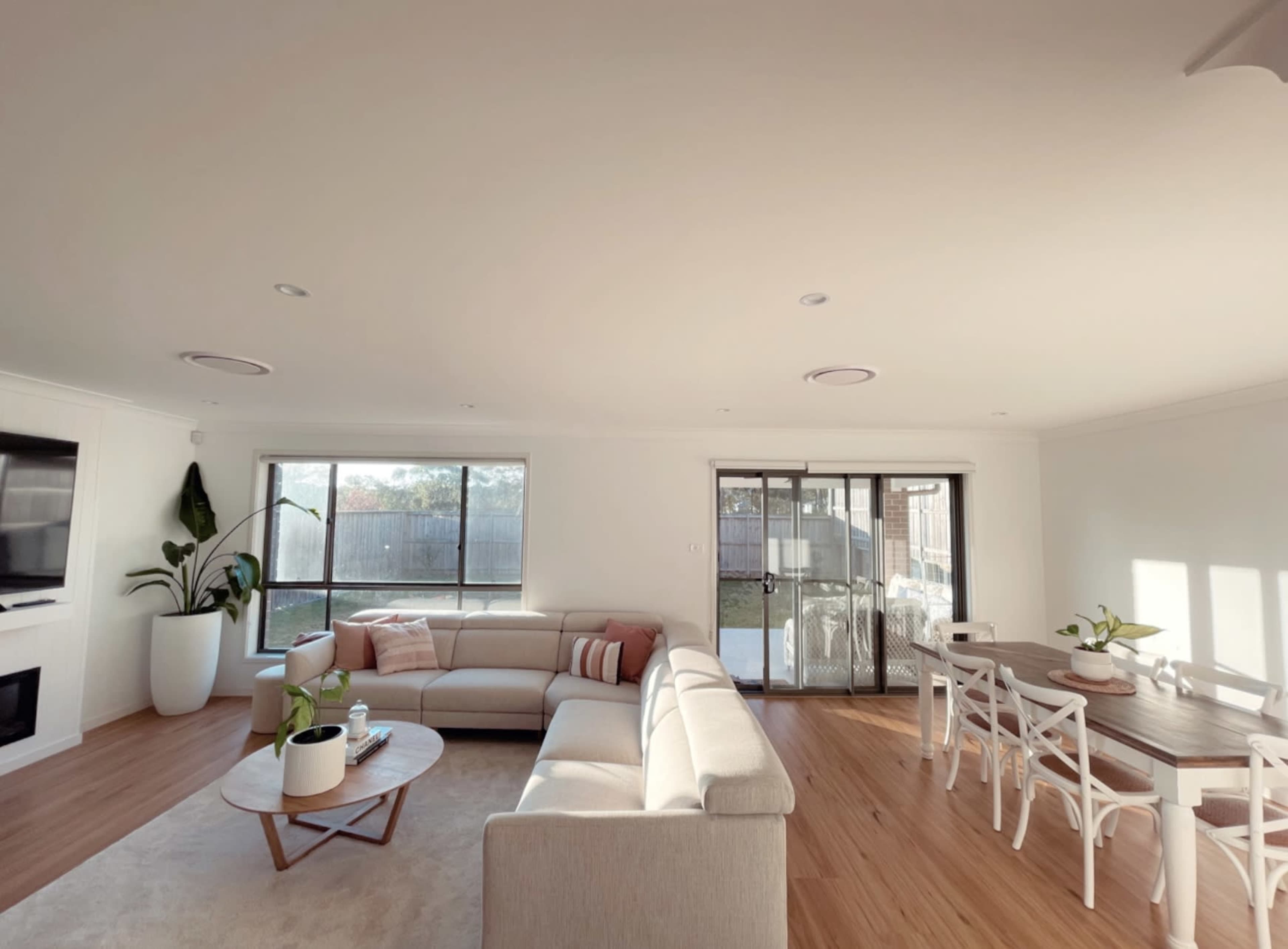 The image shows a spacious living and dining area with a light-colored sectional sofa, a circular coffee table, a dining table with chairs, and large windows overlooking an outdoor space.