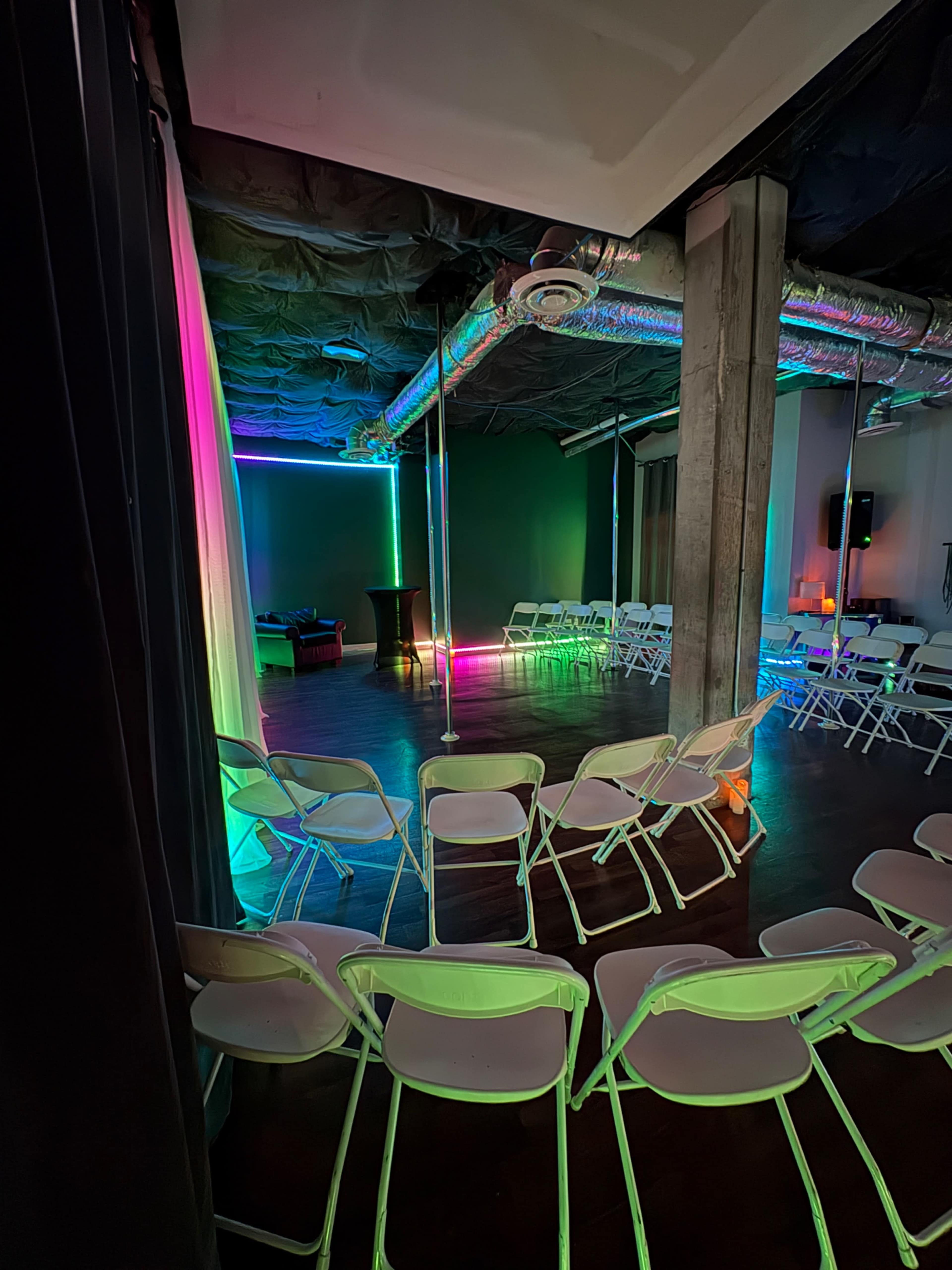 The image shows a spacious room with arranged white chairs and colorful lighting along the walls and ceiling.