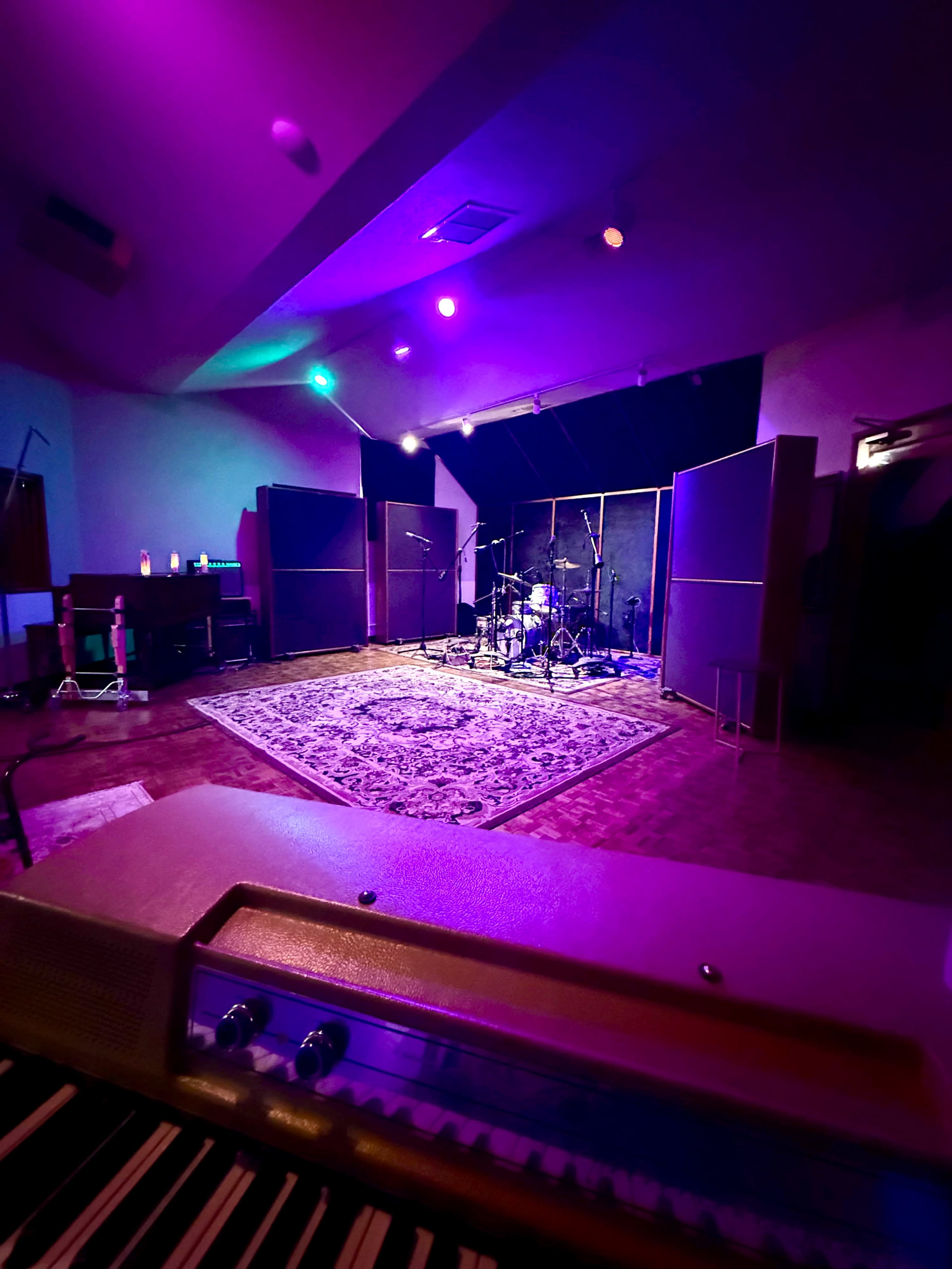 The image shows a music recording studio featuring a drum set, amplifiers, and a large area with a patterned rug under dim purple and green lighting.