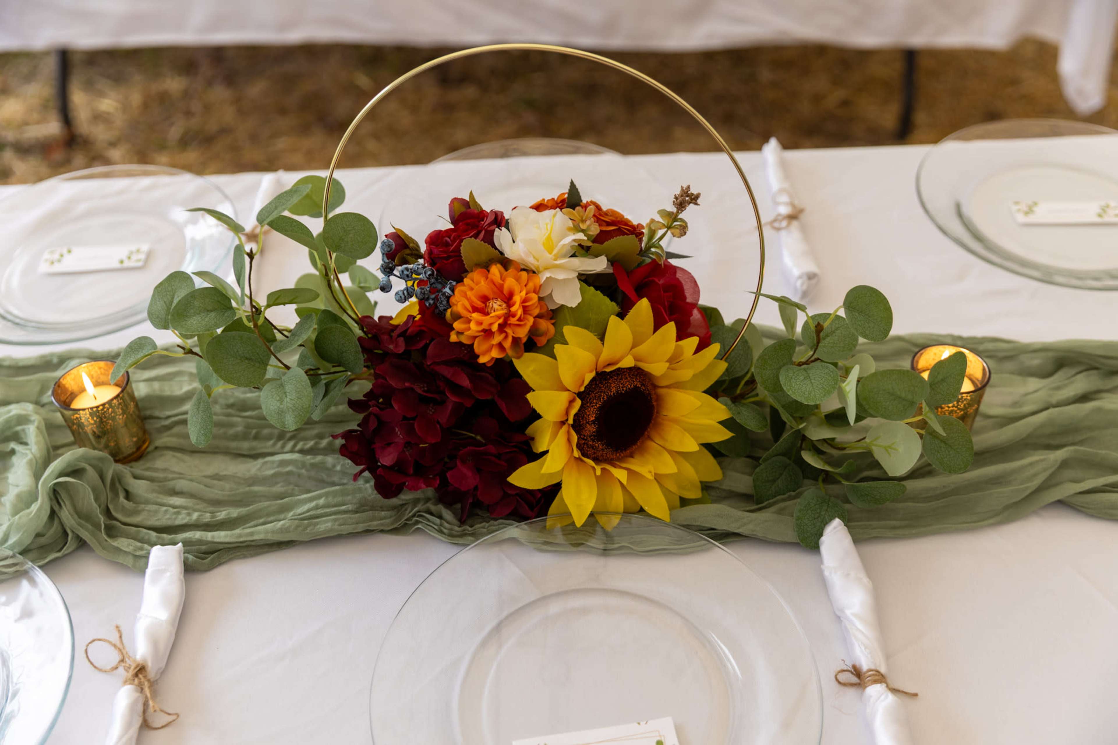 A table setting features a floral centerpiece with sunflowers, hydrangeas, and greenery, accompanied by candles, on a draped fabric runner.