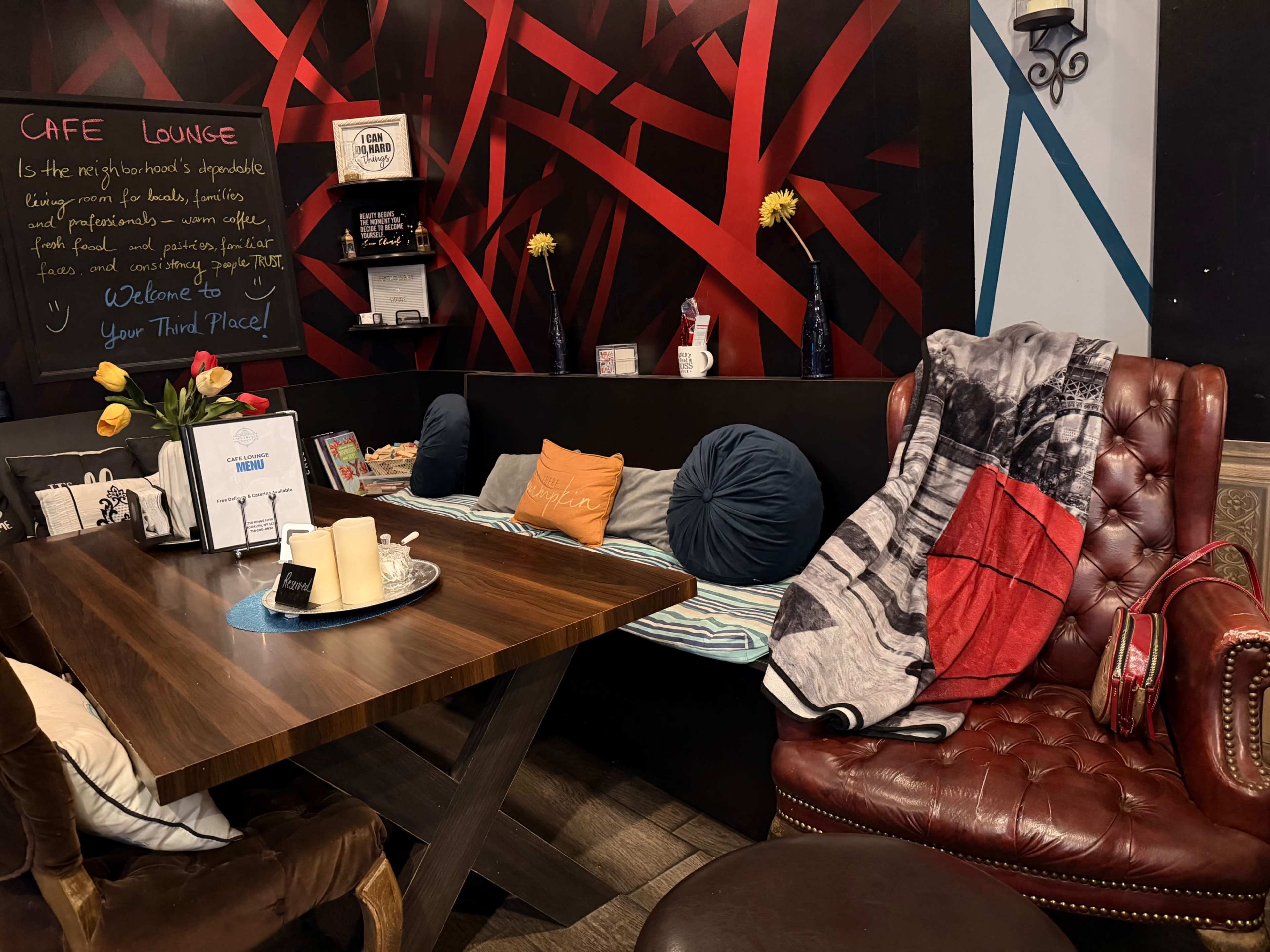 The image shows a cozy cafe lounge featuring a wooden table, various cushions, and a red and black decorative wall with a chalkboard.