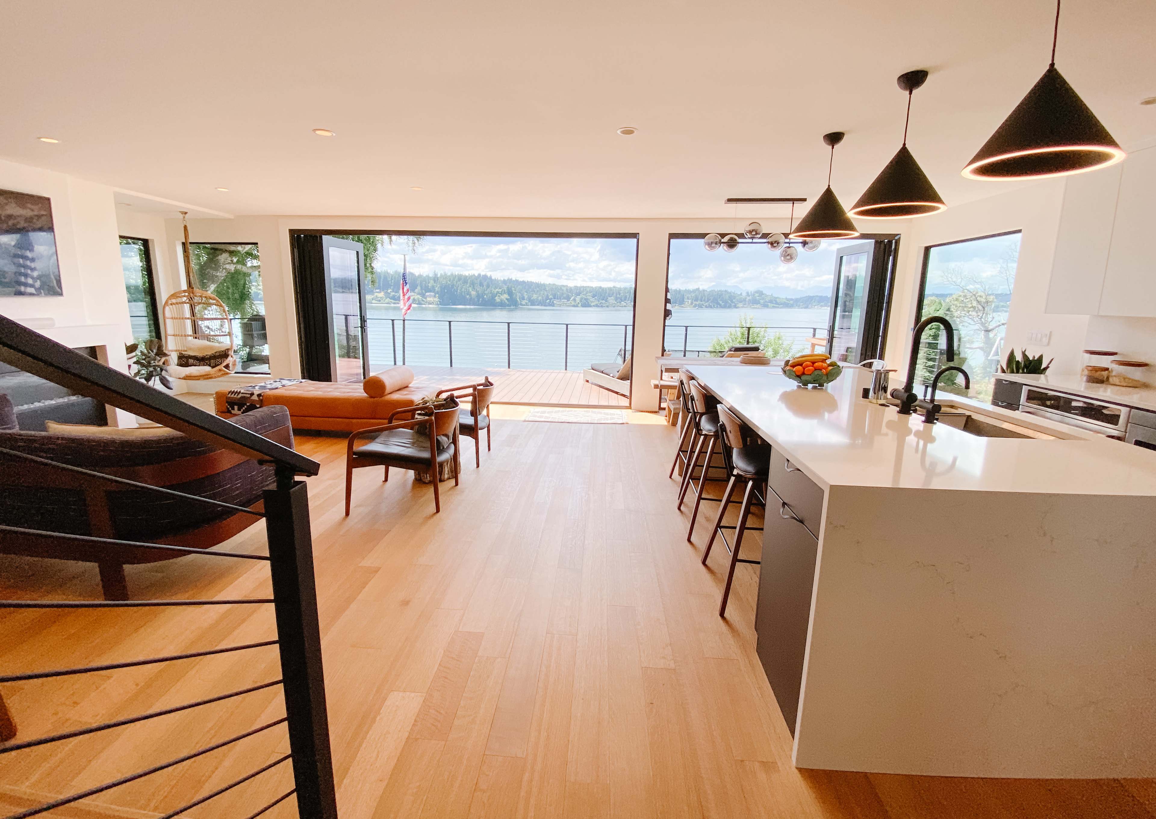 A modern, open-concept living area with a kitchen island, dining space, and large windows overlooking a body of water and deck area.