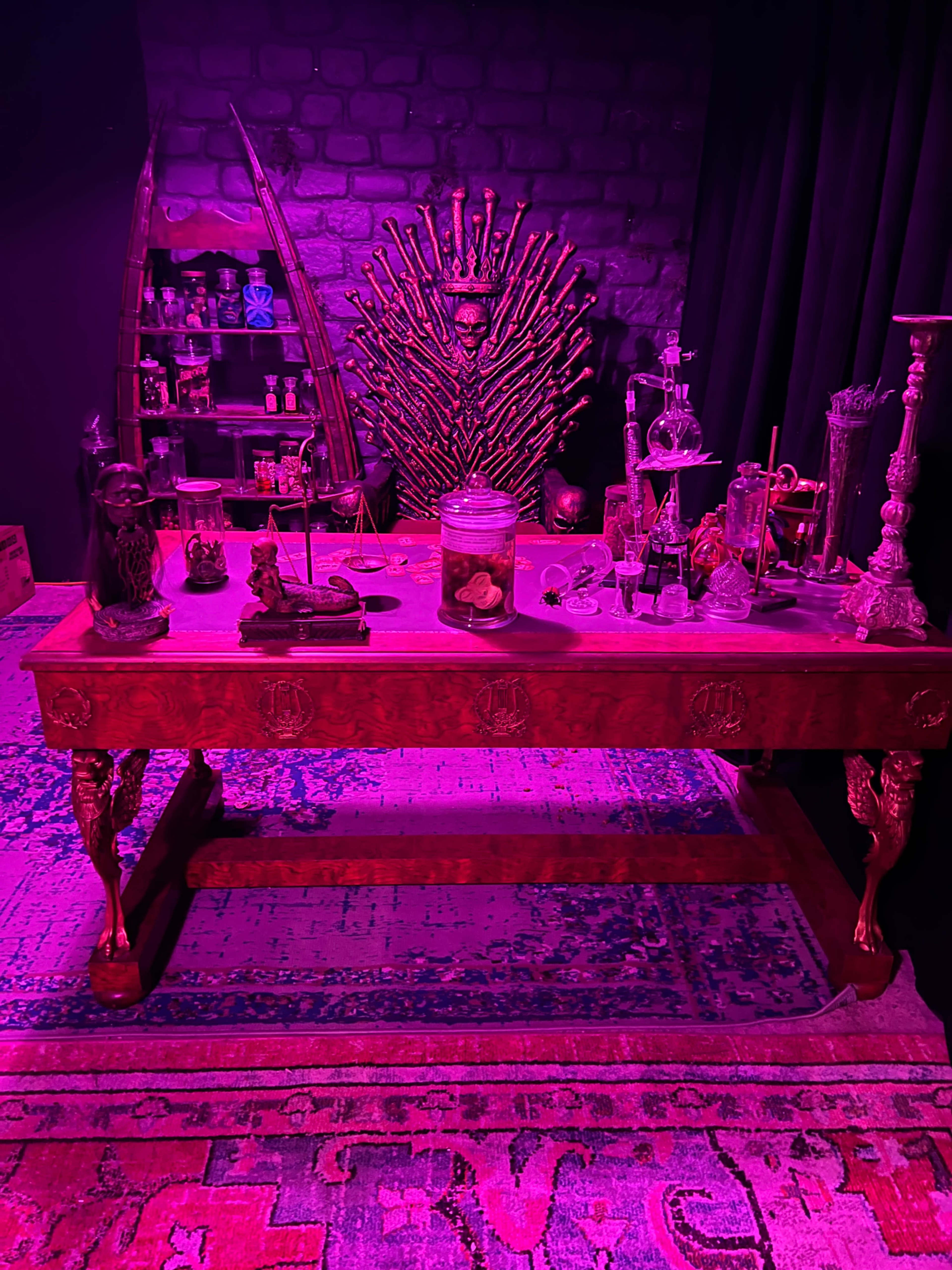 A wooden table is set with various mystical artifacts and displays under purple lighting, while a patterned rug lies beneath.