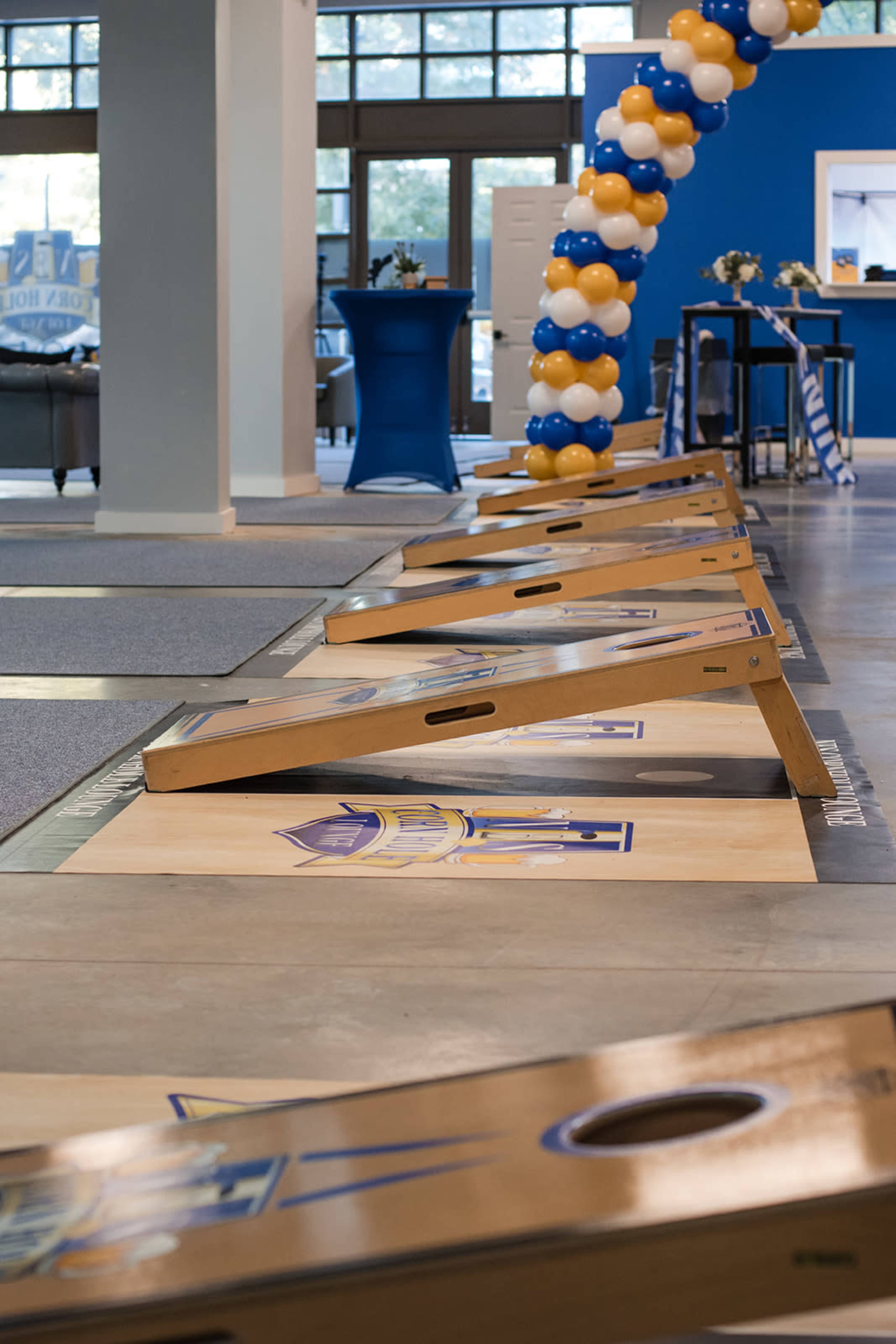The image shows a row of wooden cornhole boards set up in a spacious indoor area decorated with blue and gold balloons.