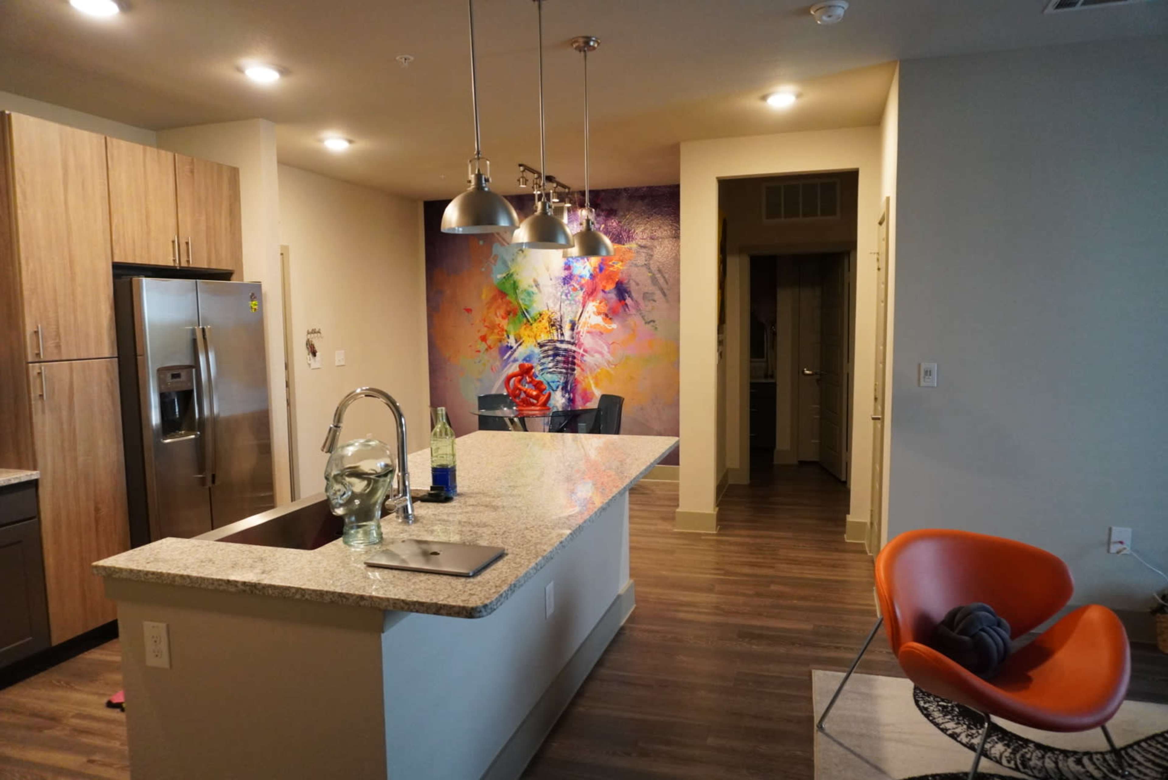 A modern kitchen with granite countertops, stainless steel appliances, and a vibrant mural on the wall, leading to a hallway with an orange chair nearby.