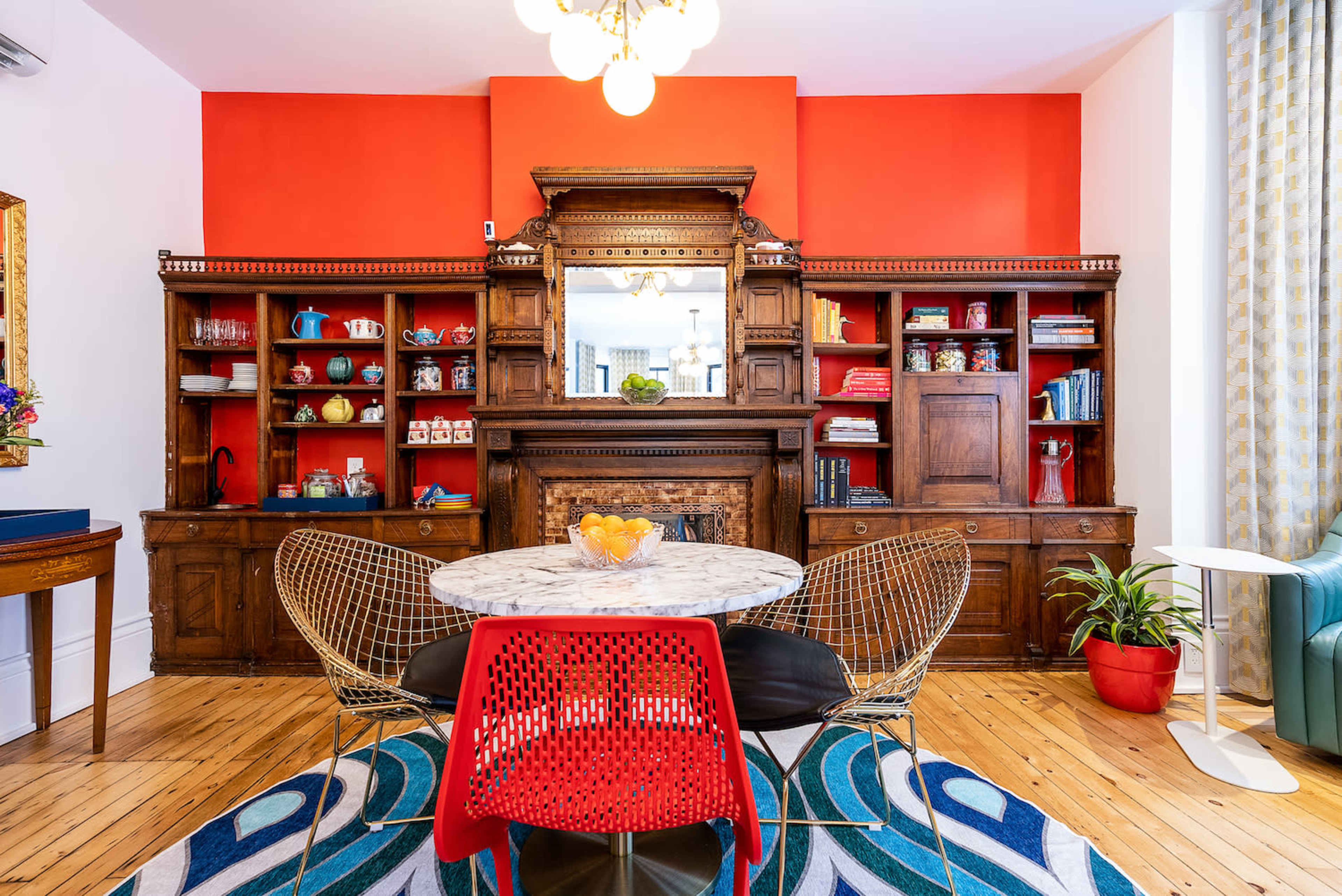 A dining area features a round marble table surrounded by colorful chairs, set against a bright red wall with an ornate wooden cabinet displaying various dishes and books.