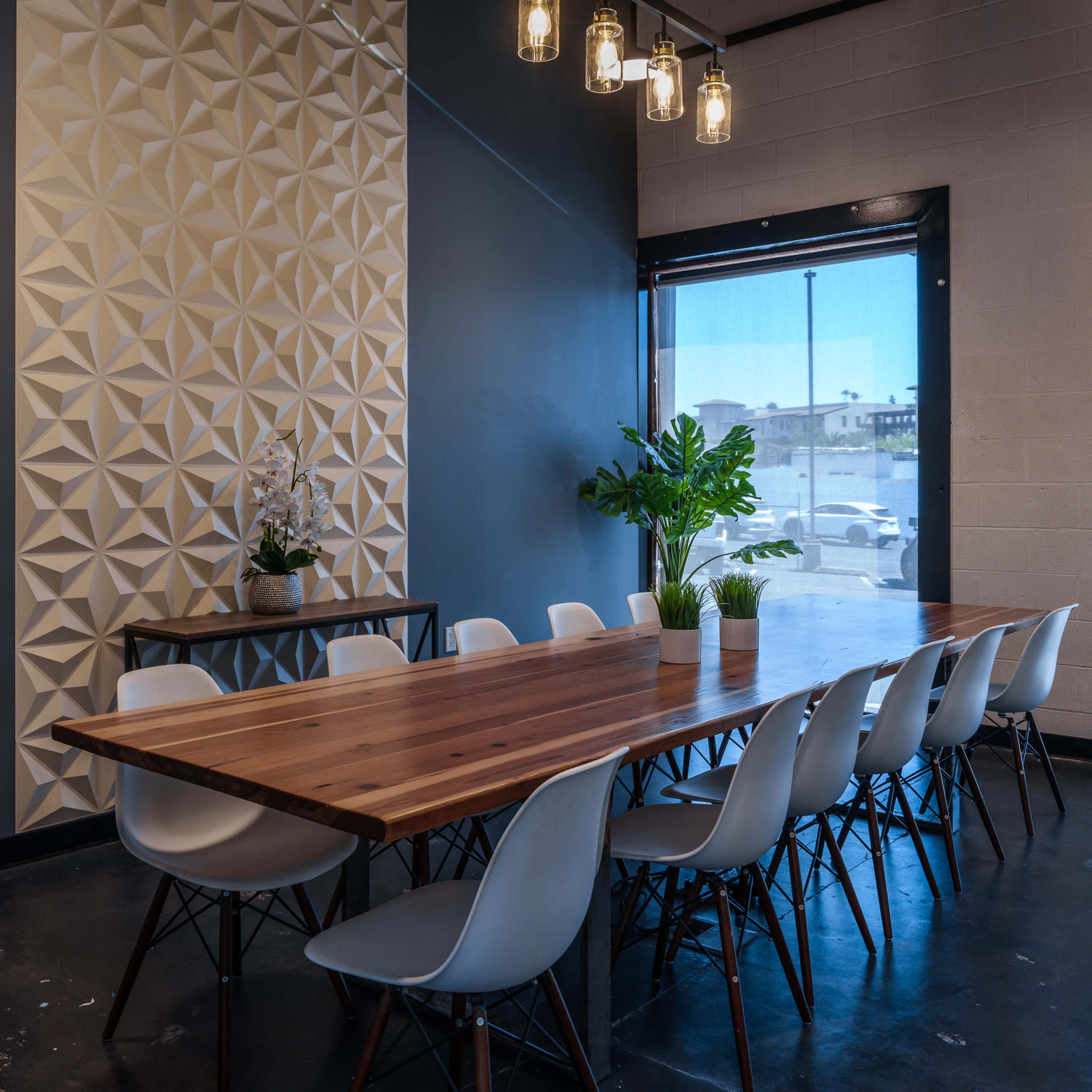 A long wooden dining table with white chairs is set under pendant lights in a room featuring a textured wall and a large window.
