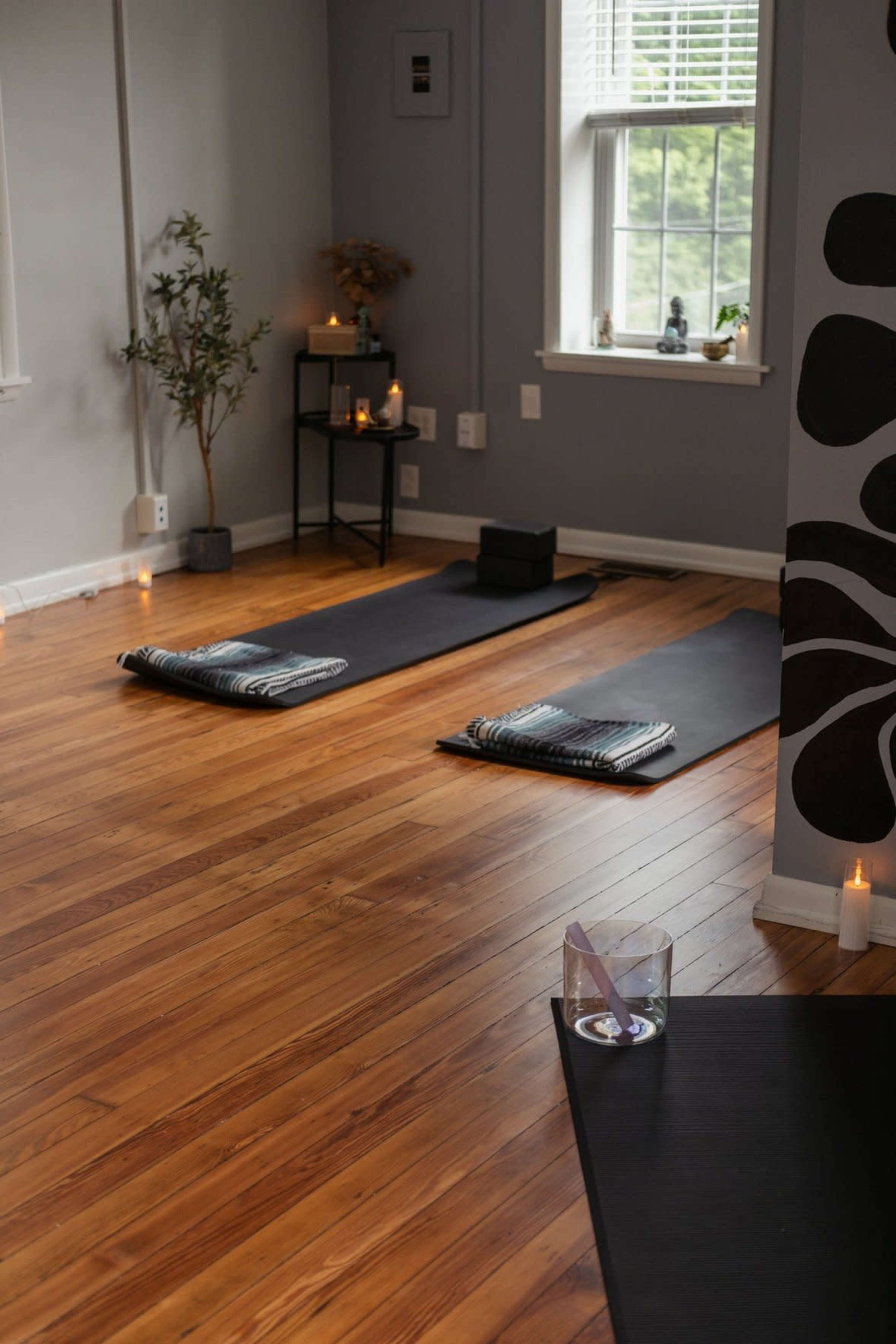 The image shows a calm yoga studio featuring two yoga mats on a wooden floor, a small plant, a shelf, candles, and a decorative wall.