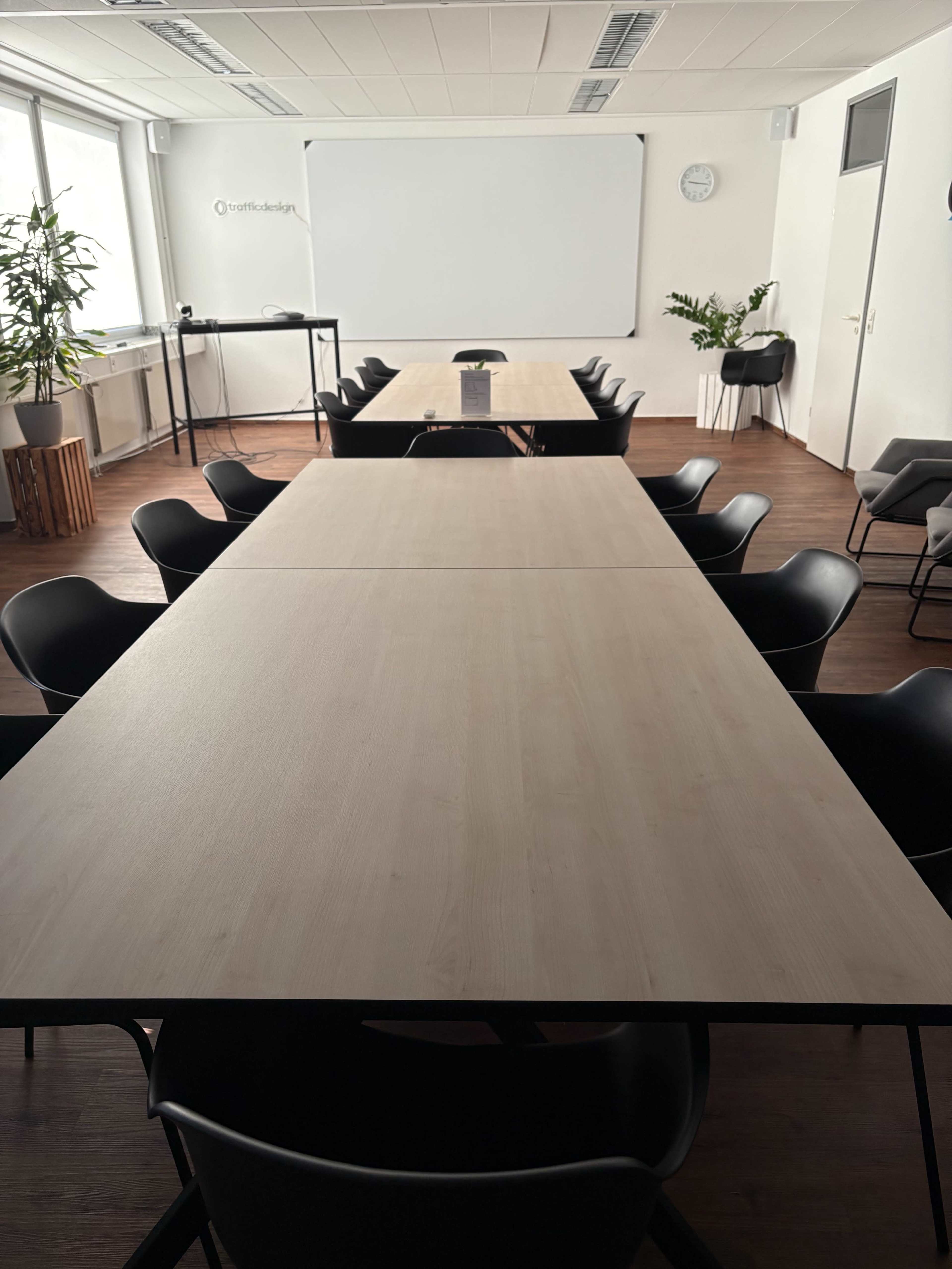 The image depicts a spacious conference room with long tables arranged in a rectangular layout and black chairs surrounding them.
