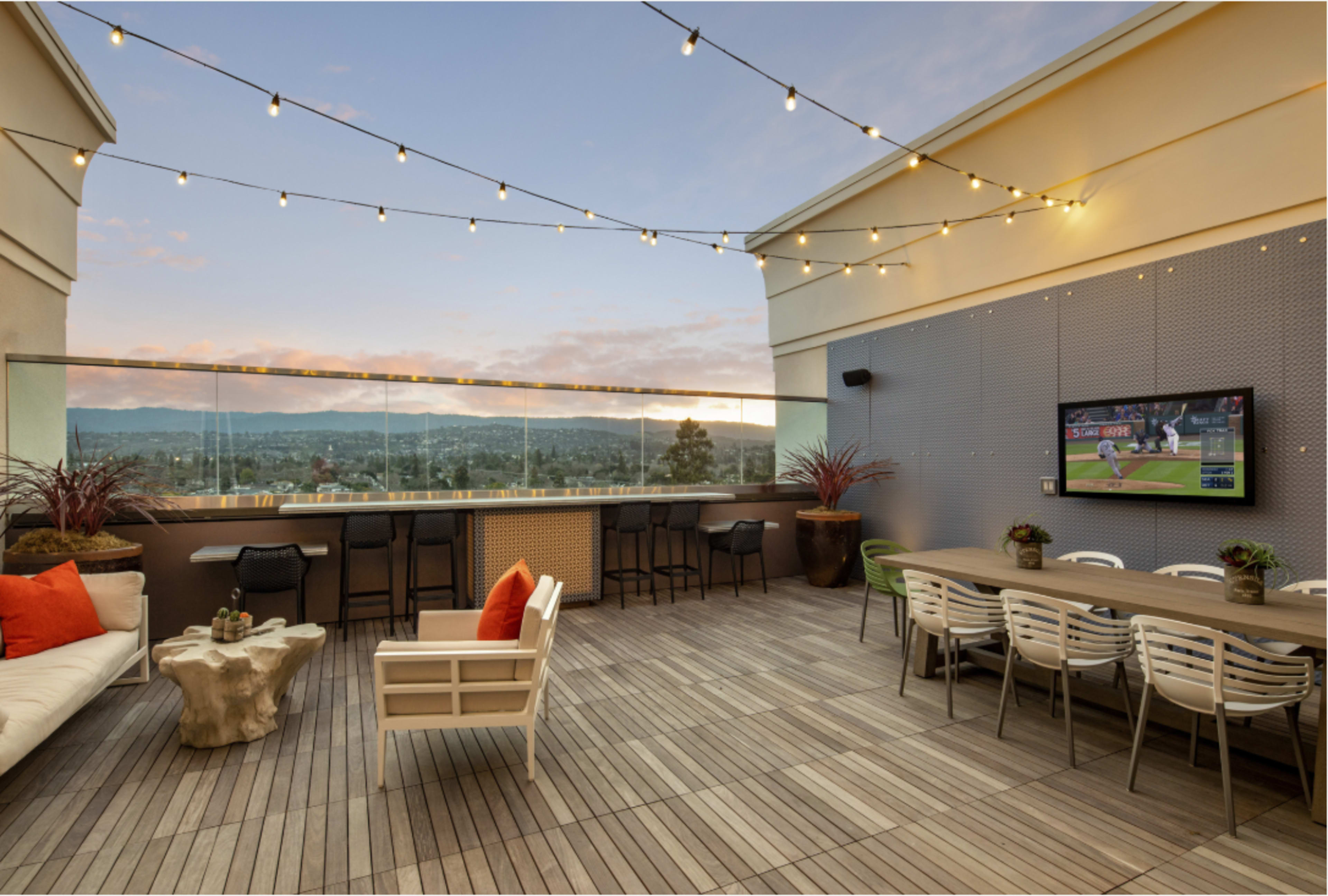A rooftop terrace features a table with chairs, a sofa, and a large television, surrounded by string lights and a scenic view of the landscape.