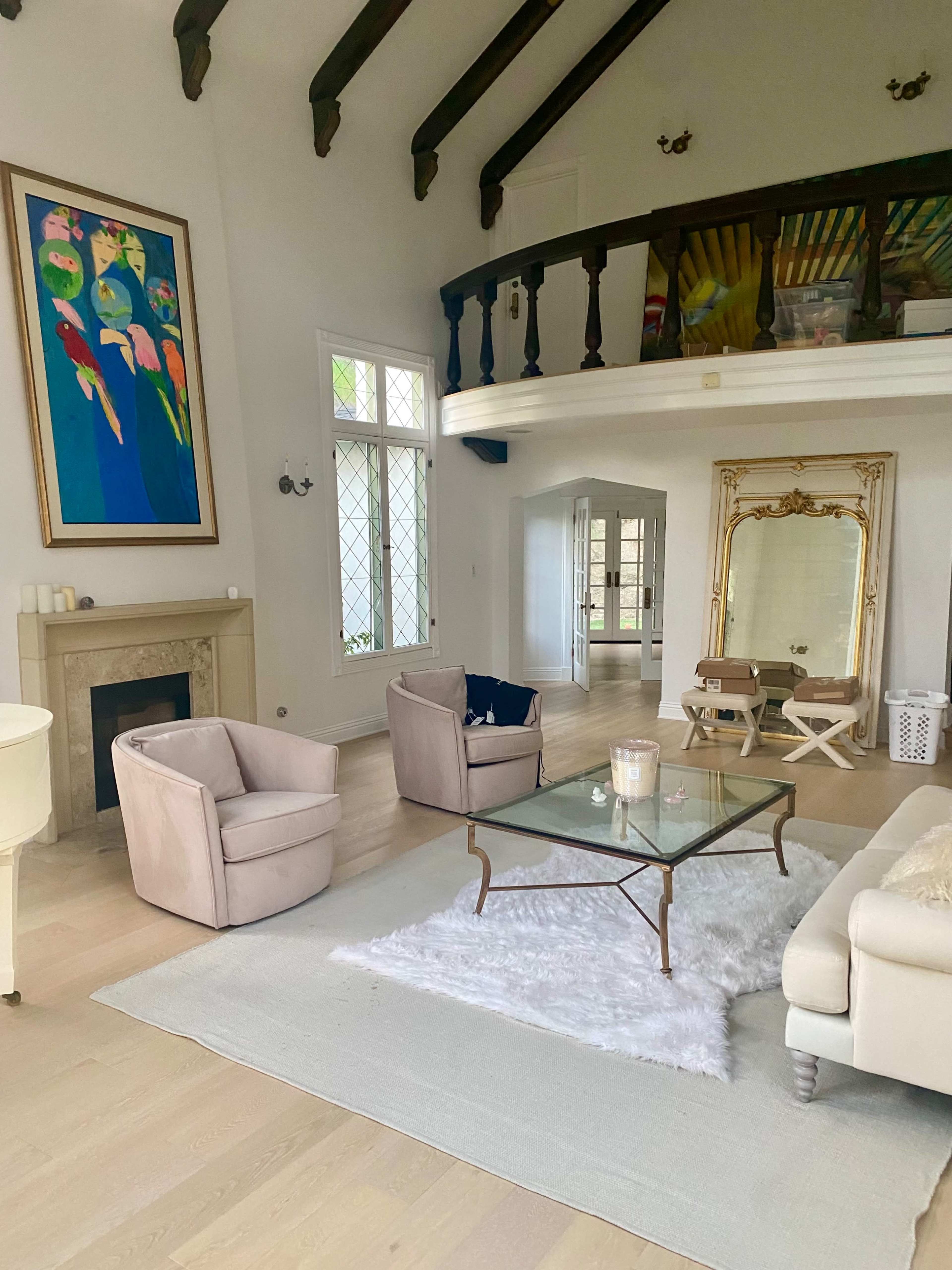 A spacious living room featuring a high ceiling, a glass coffee table on a white rug, two pale armchairs, and a large artwork of colorful birds on the wall.