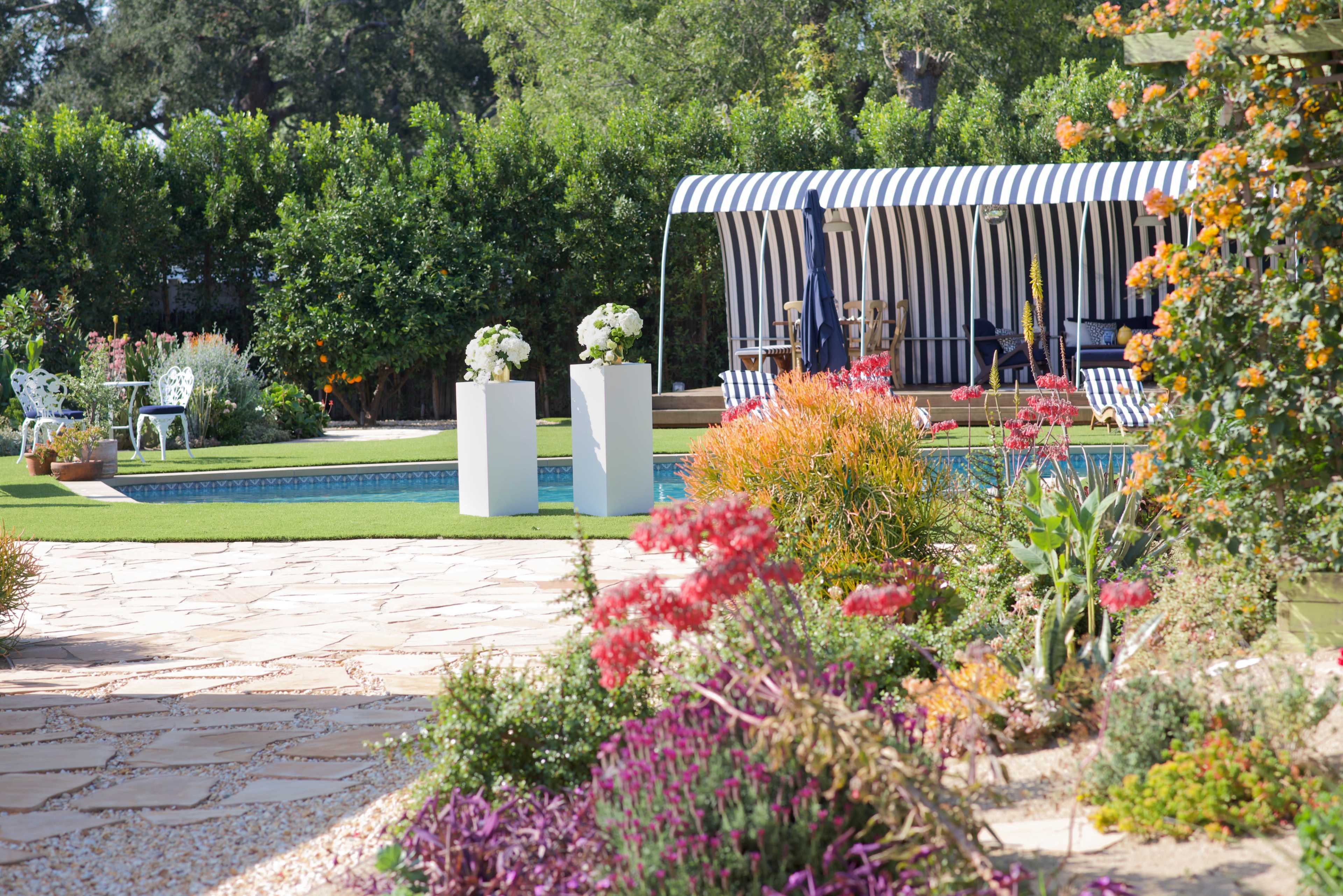 The image shows a landscaped garden with colorful flowering plants, a swimming pool, and a striped cabana in the background.