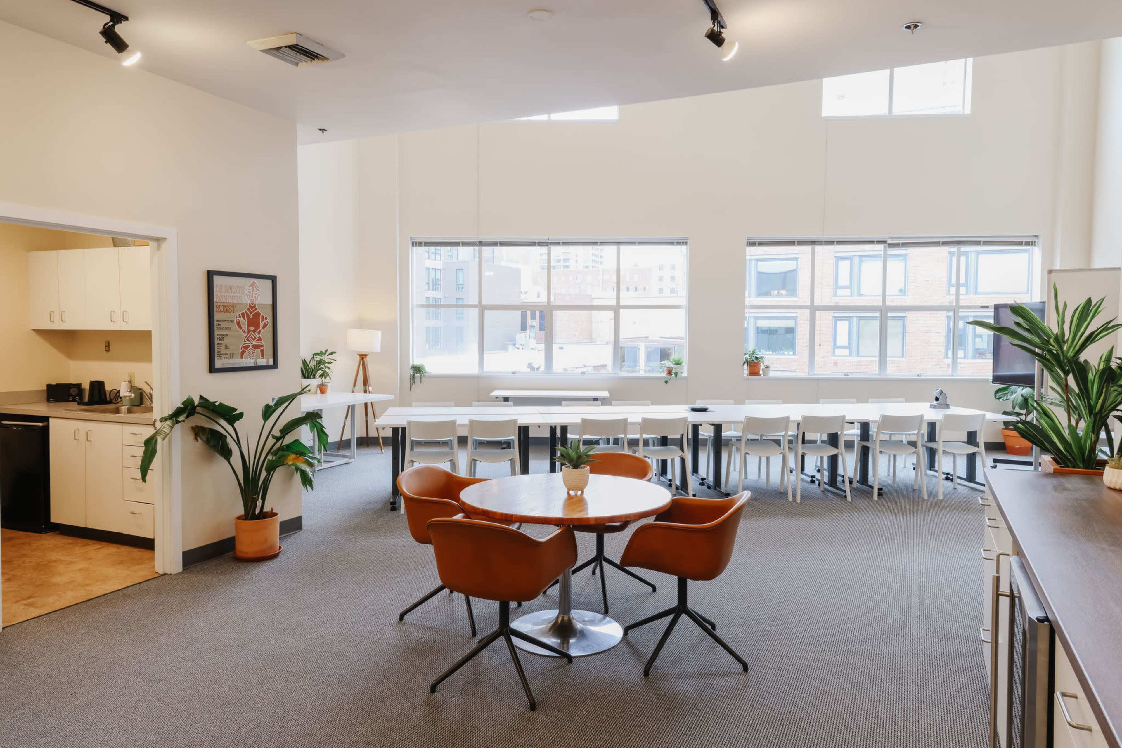 The image shows a spacious, well-lit room with a dining area featuring a round table and chairs, surrounded by large windows and kitchen facilities.