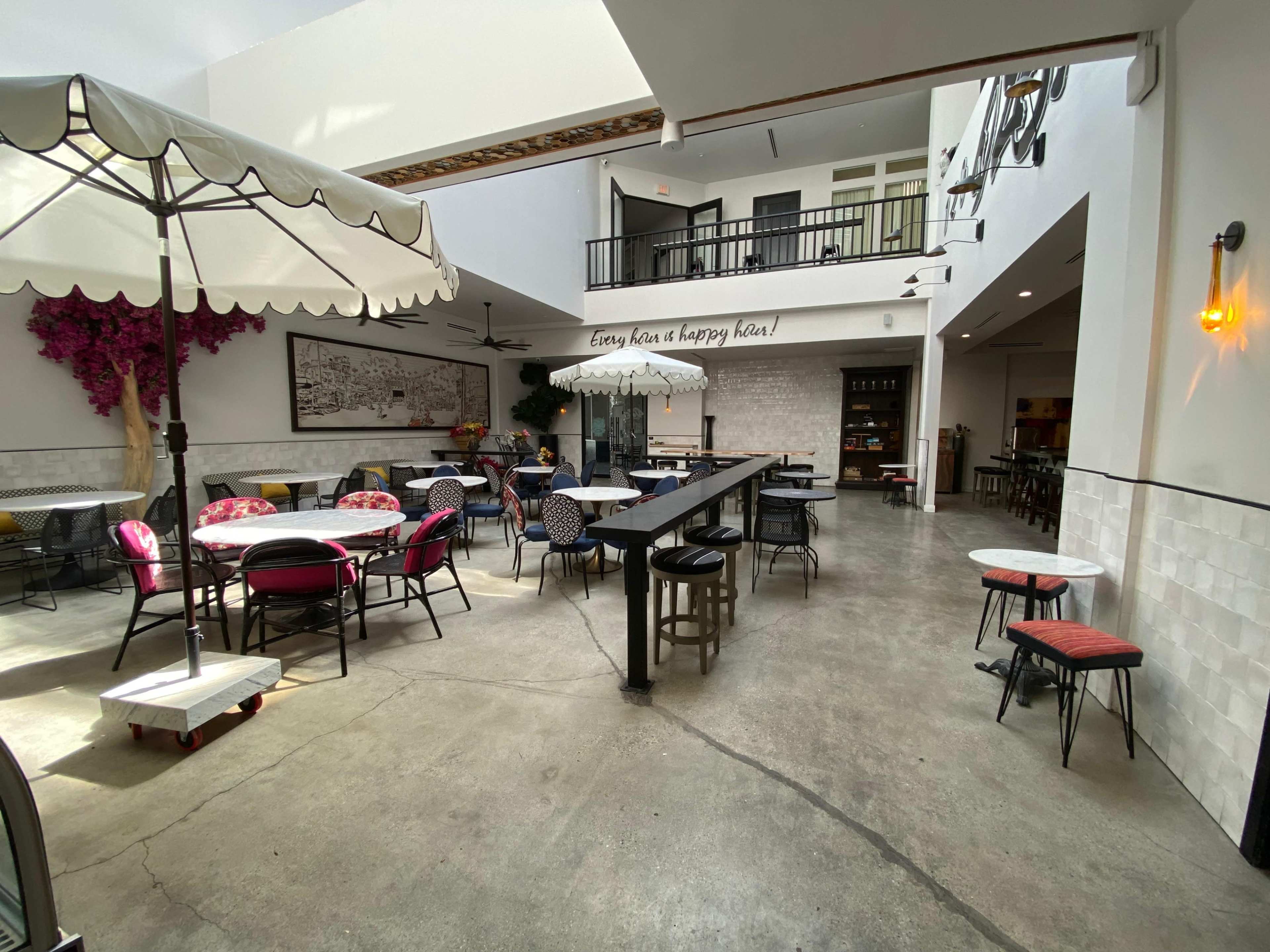 The image shows a spacious, modern indoor dining area featuring a mix of tables and chairs, with a large umbrella and decorative wall plants.