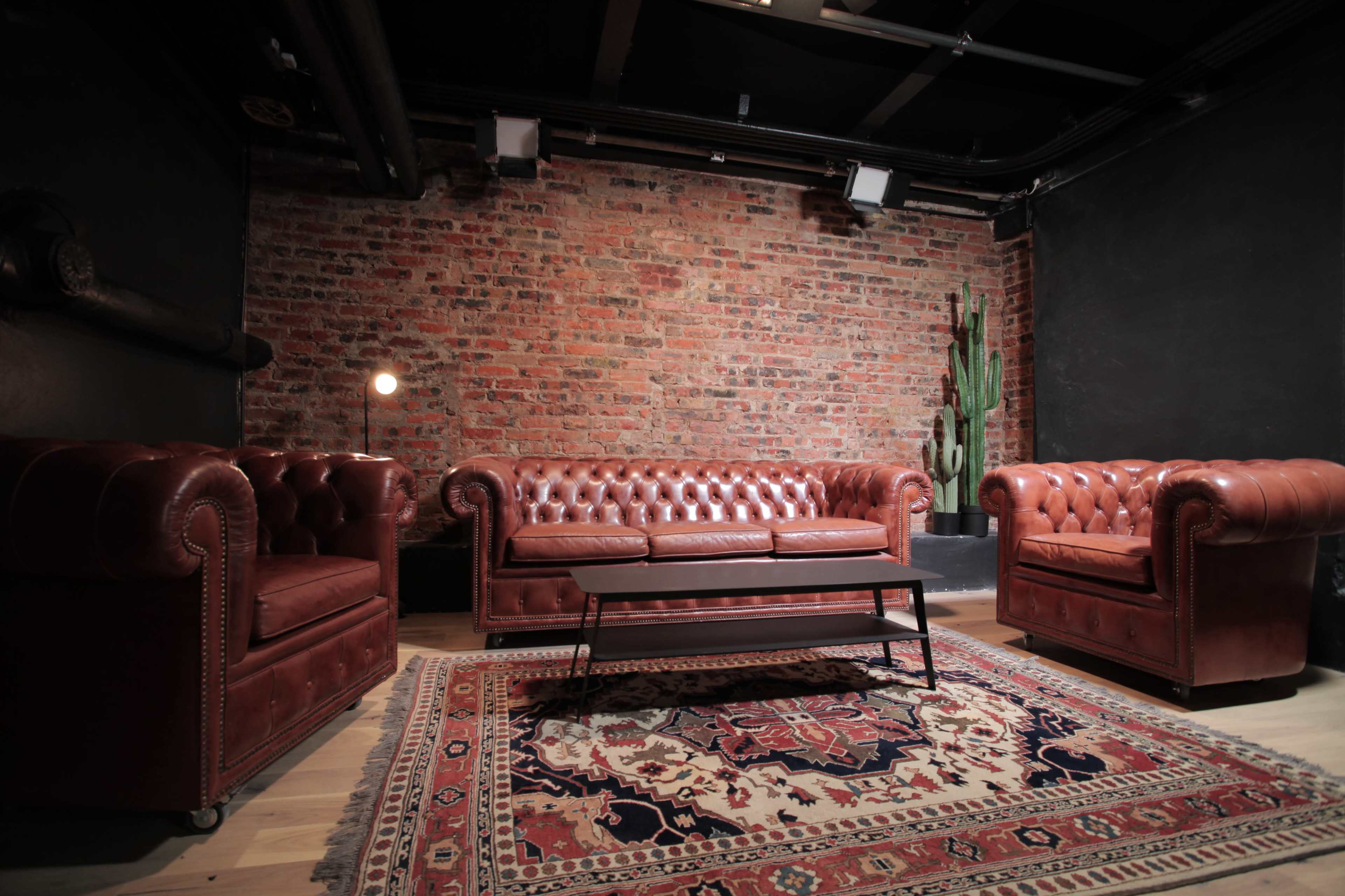 The image depicts a cozy seating area furnished with brown leather chesterfield sofas and a coffee table on a patterned rug, set against a brick wall with a cactus in the corner.