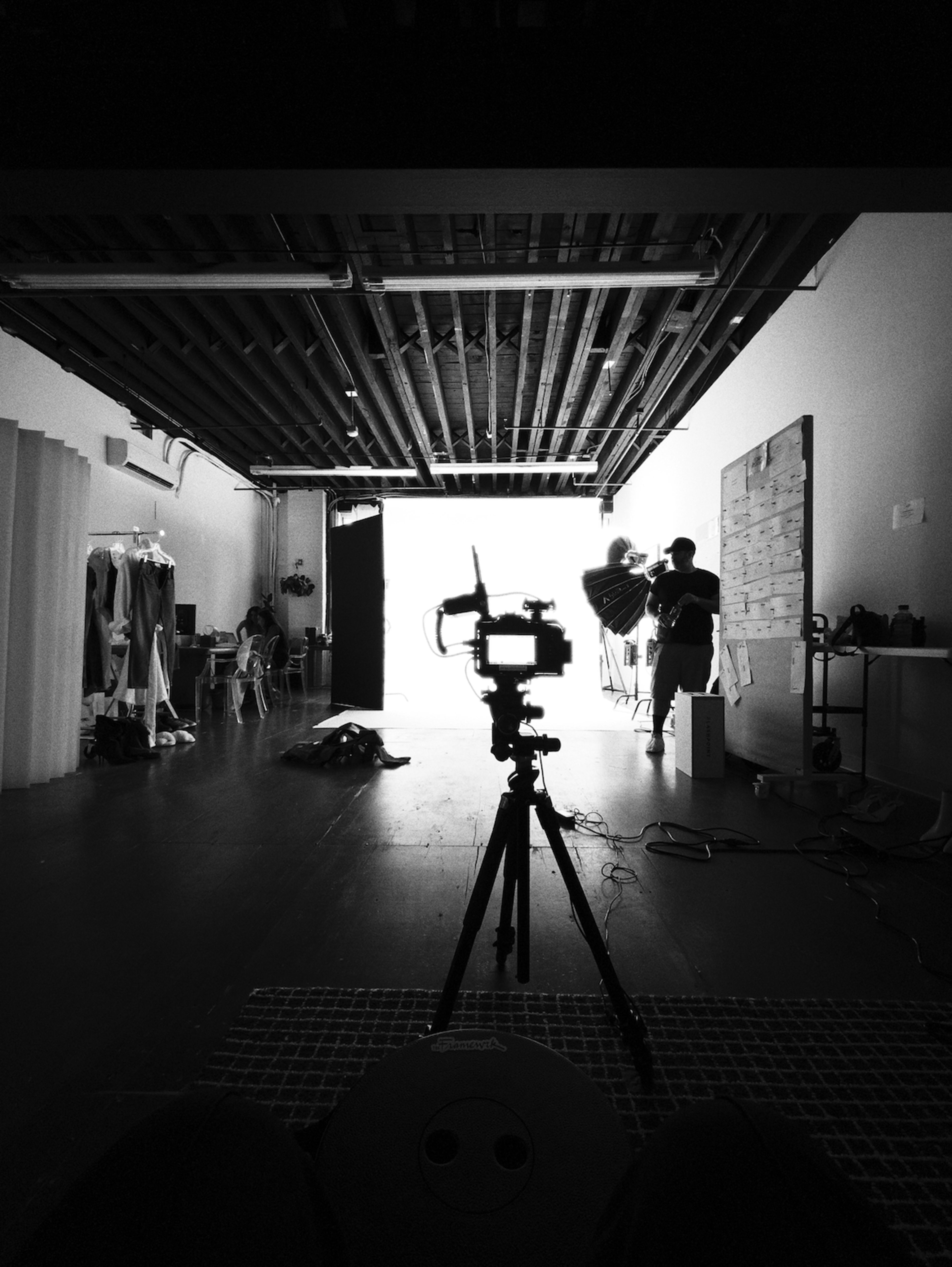 A camera on a tripod is pointed toward a brightly lit doorway in a spacious photography studio, with equipment and clothing racks visible around the room.