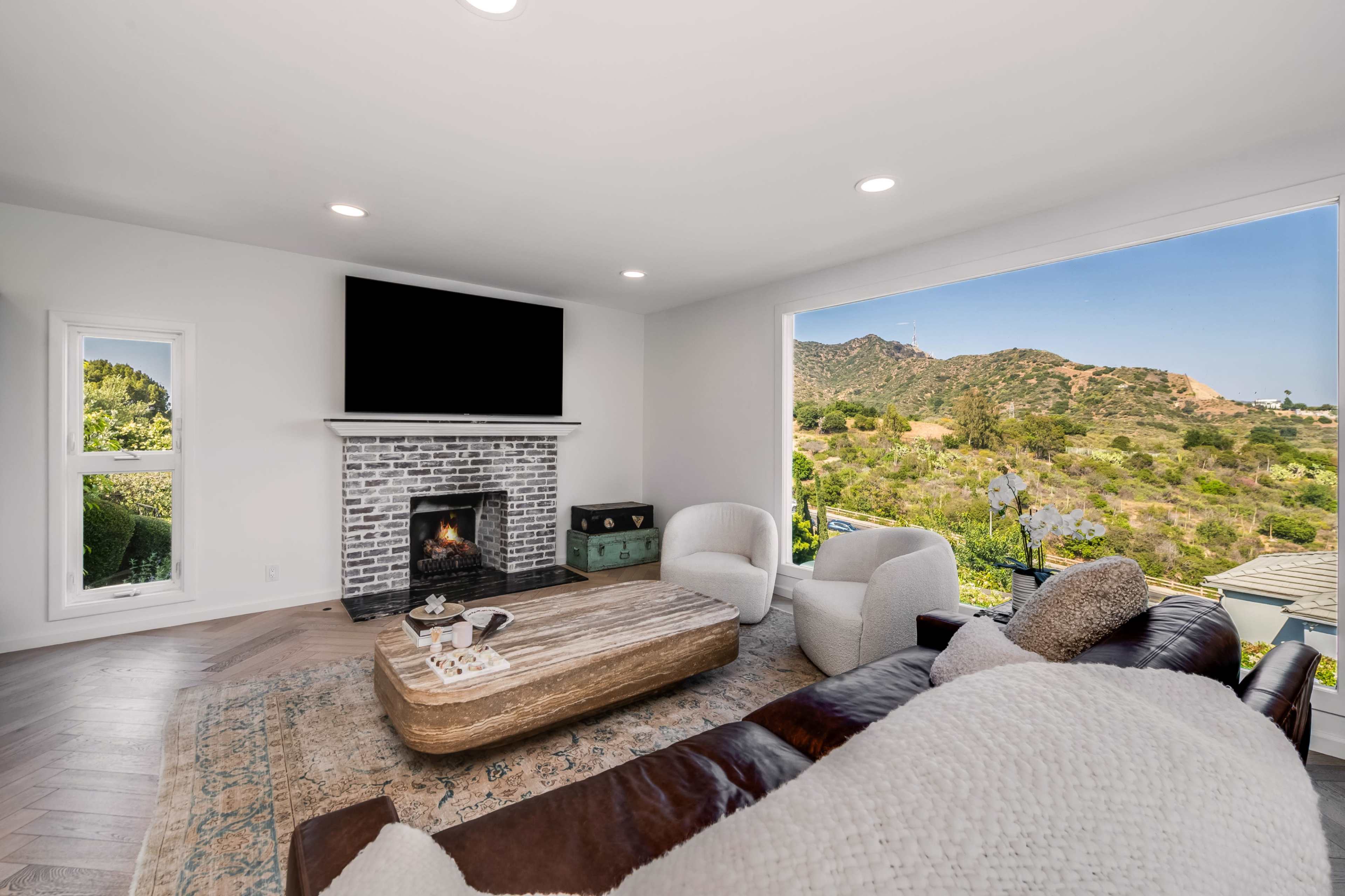 A modern living room features a large window overlooking a hilly landscape, a fireplace with a brick facade, and a wooden coffee table surrounded by comfortable seating.