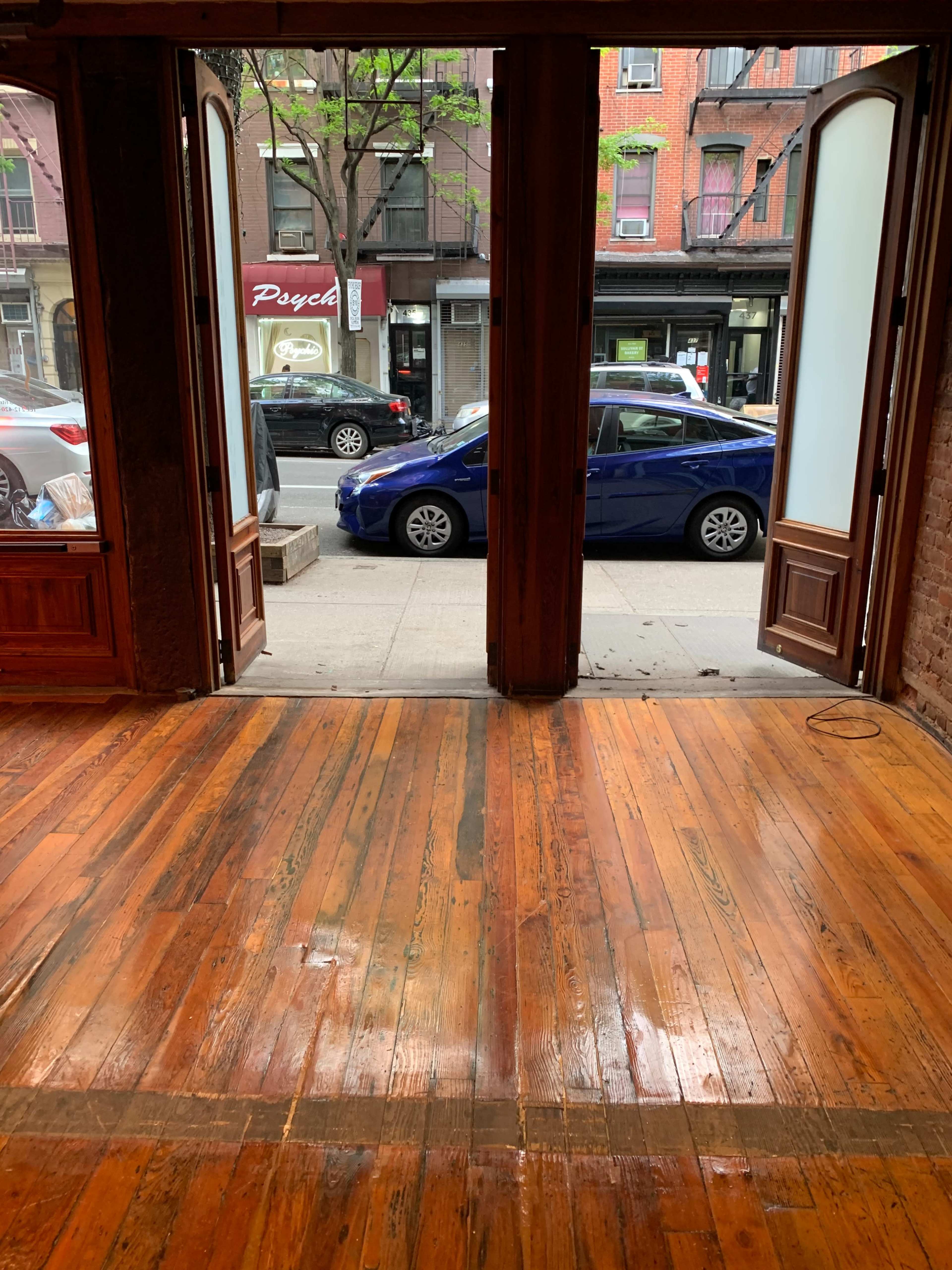 An empty interior with polished wooden floors facing an open doorway that leads to a street with parked cars and a storefront.
