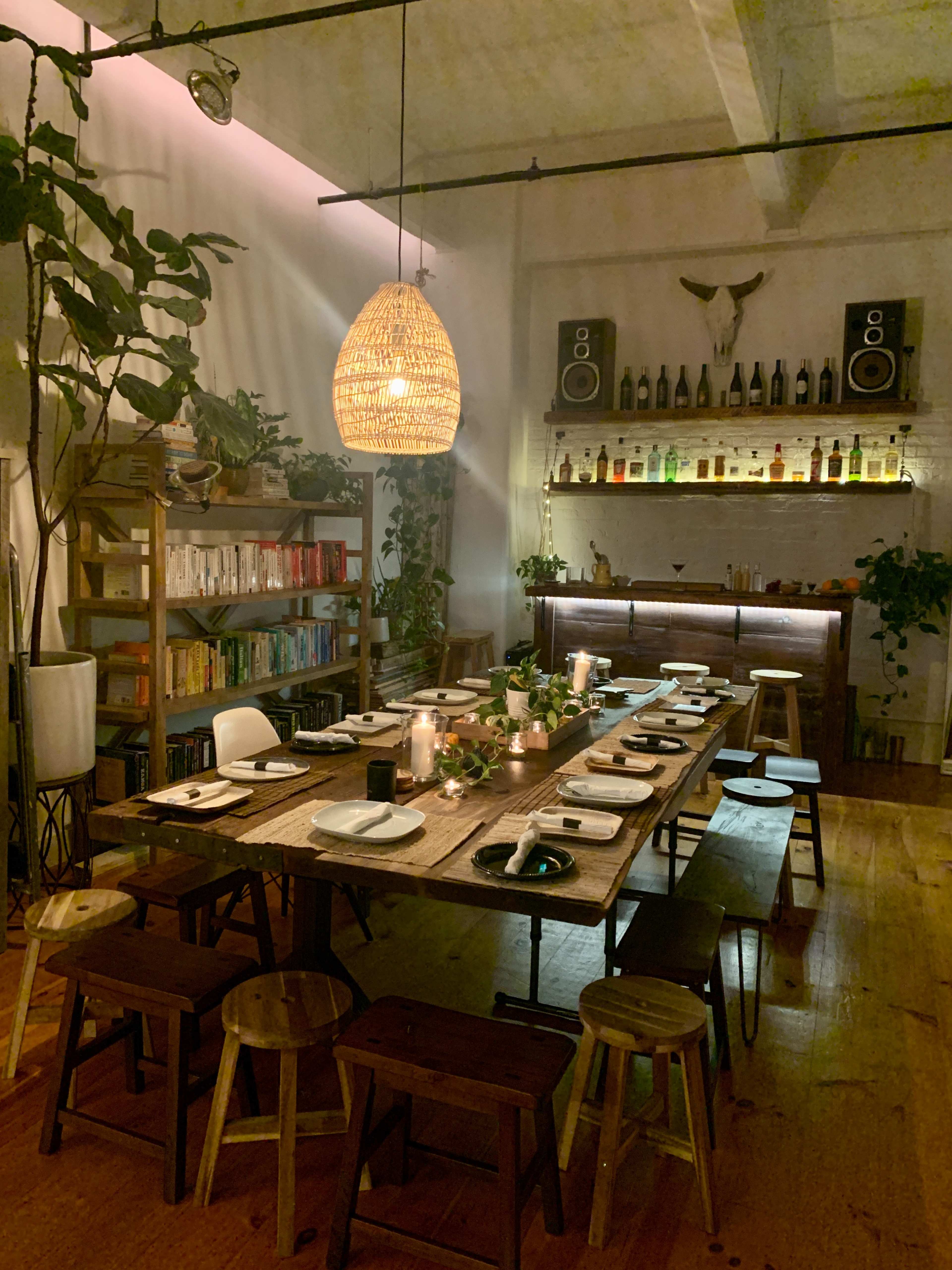 A long communal dining table is set up in a dimly lit room adorned with plants, shelves of books, and a bar in the background.