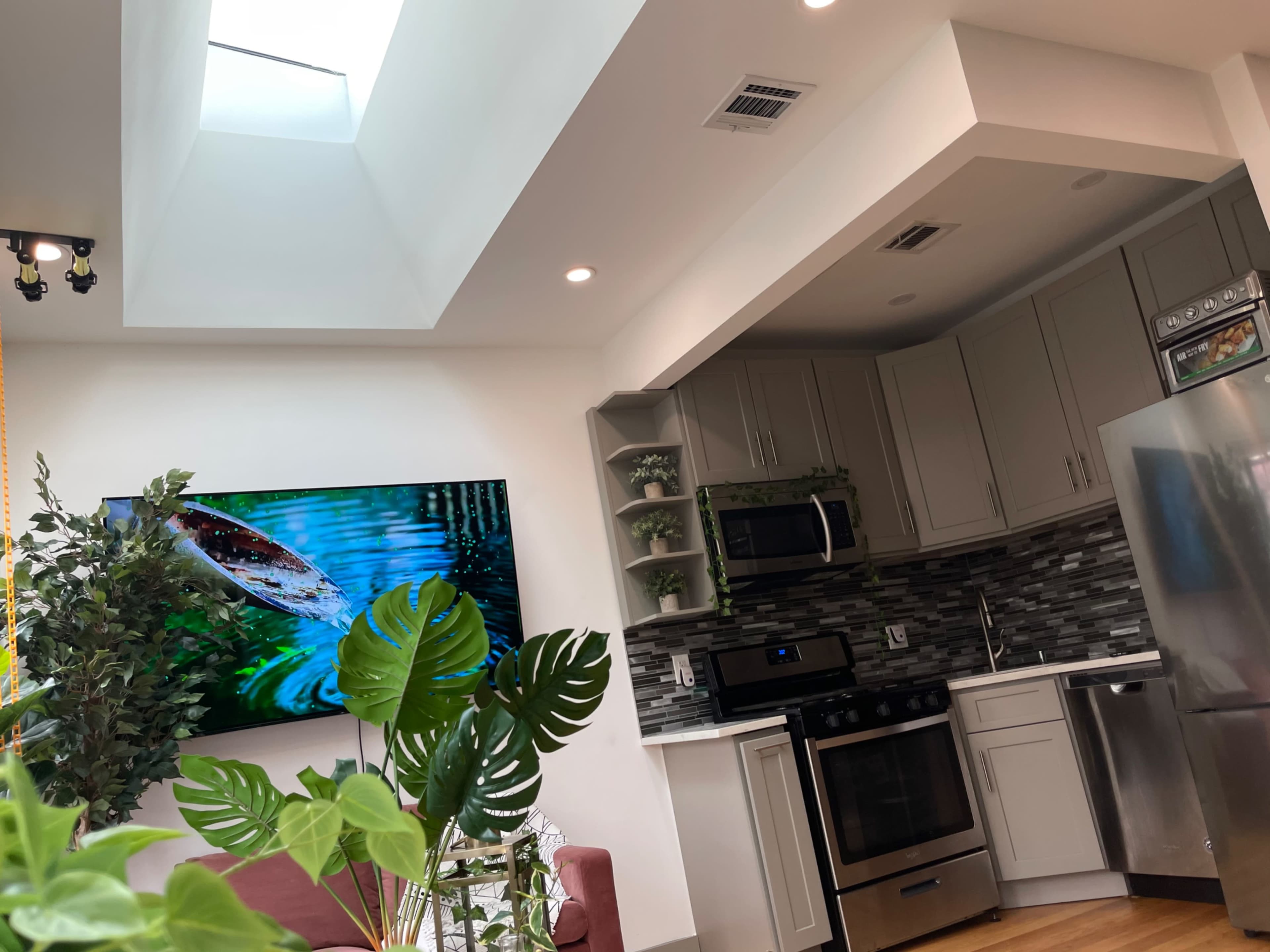 A modern kitchen and living area featuring a skylight, stainless steel appliances, and a large television mounted on the wall.
