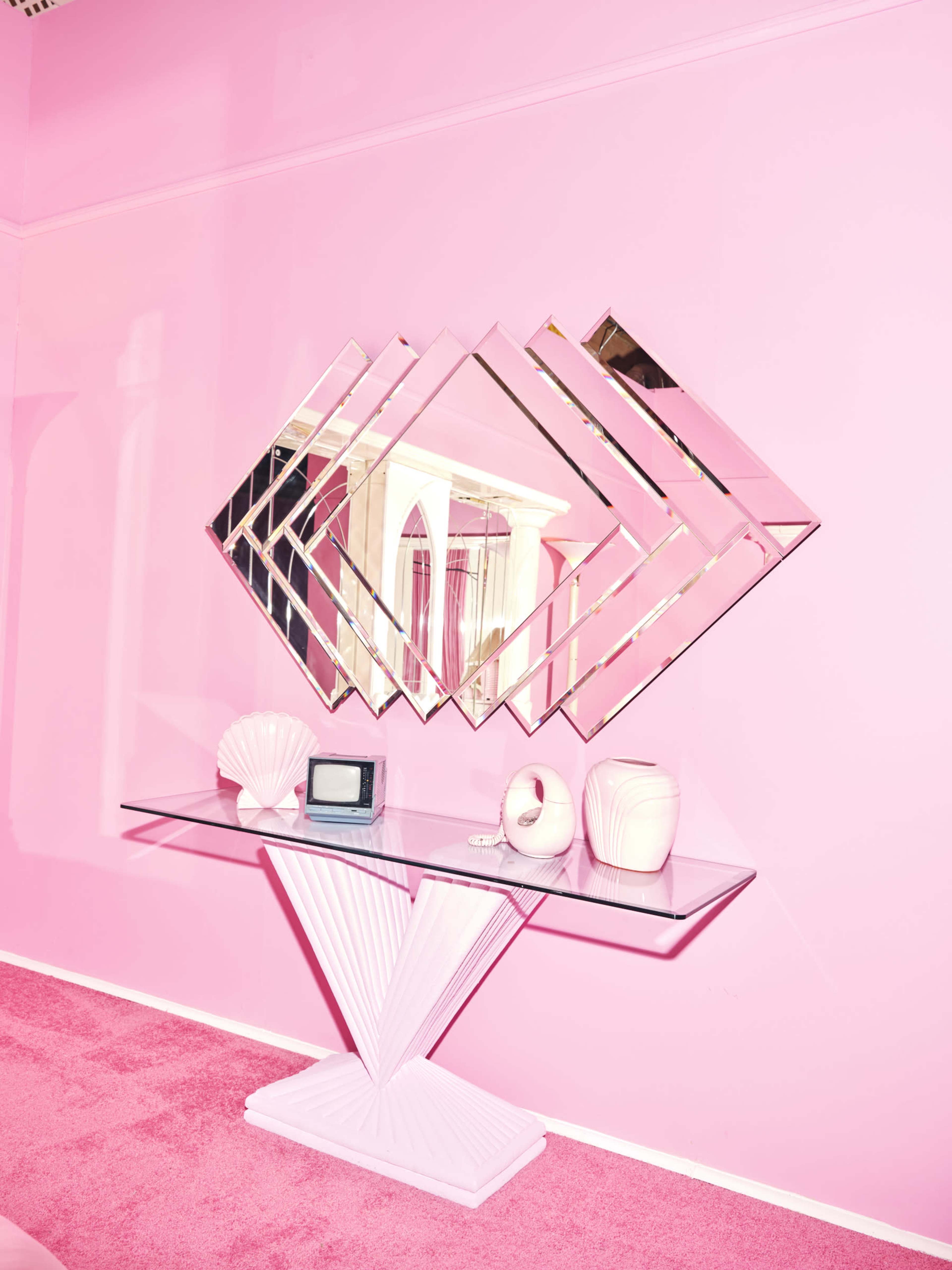 A mirrored geometric decoration is mounted on a pink wall above a glass table with a white shell, a vintage television, and two vases.