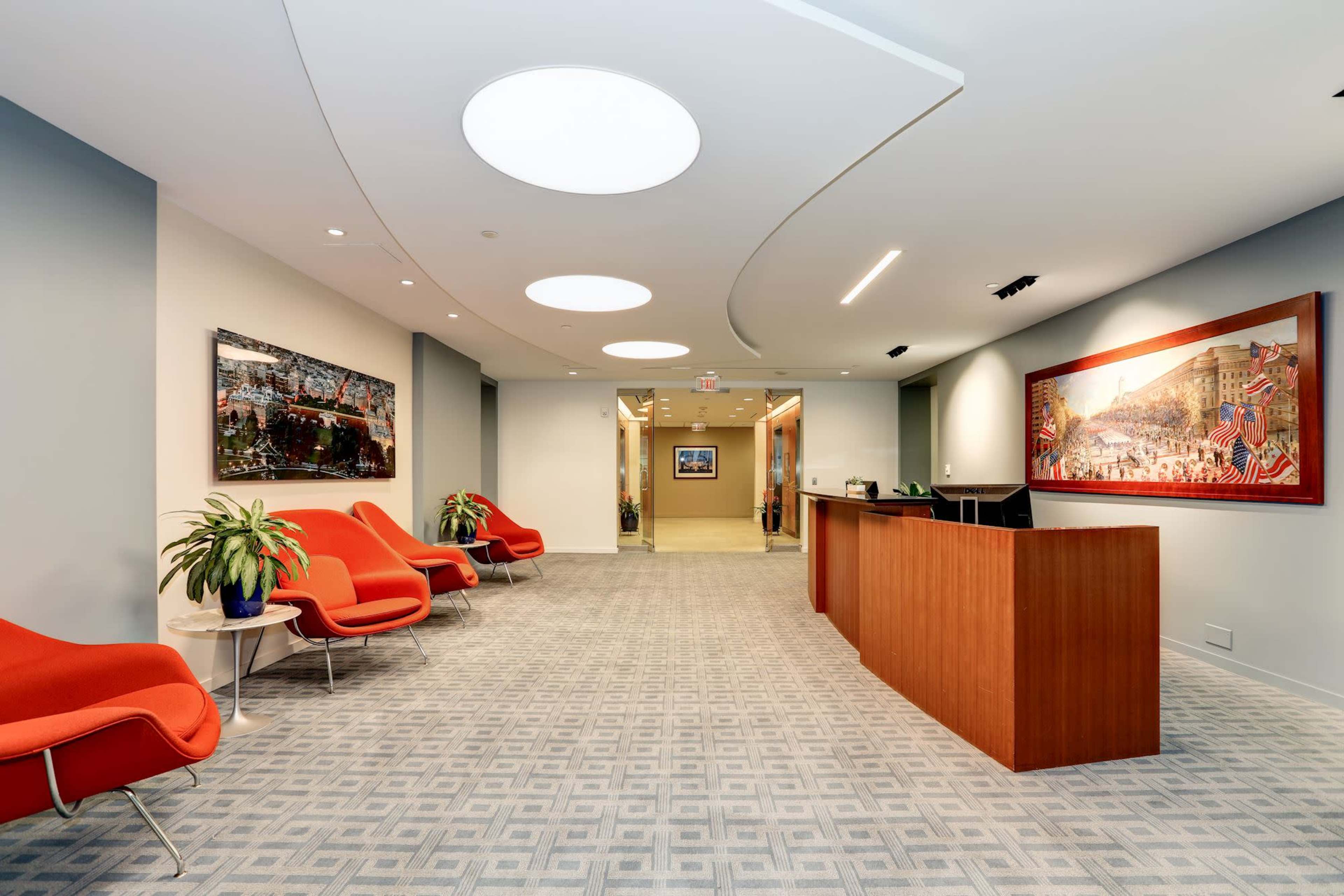 A modern office reception area with a wooden front desk, red chairs, and artwork on the walls.