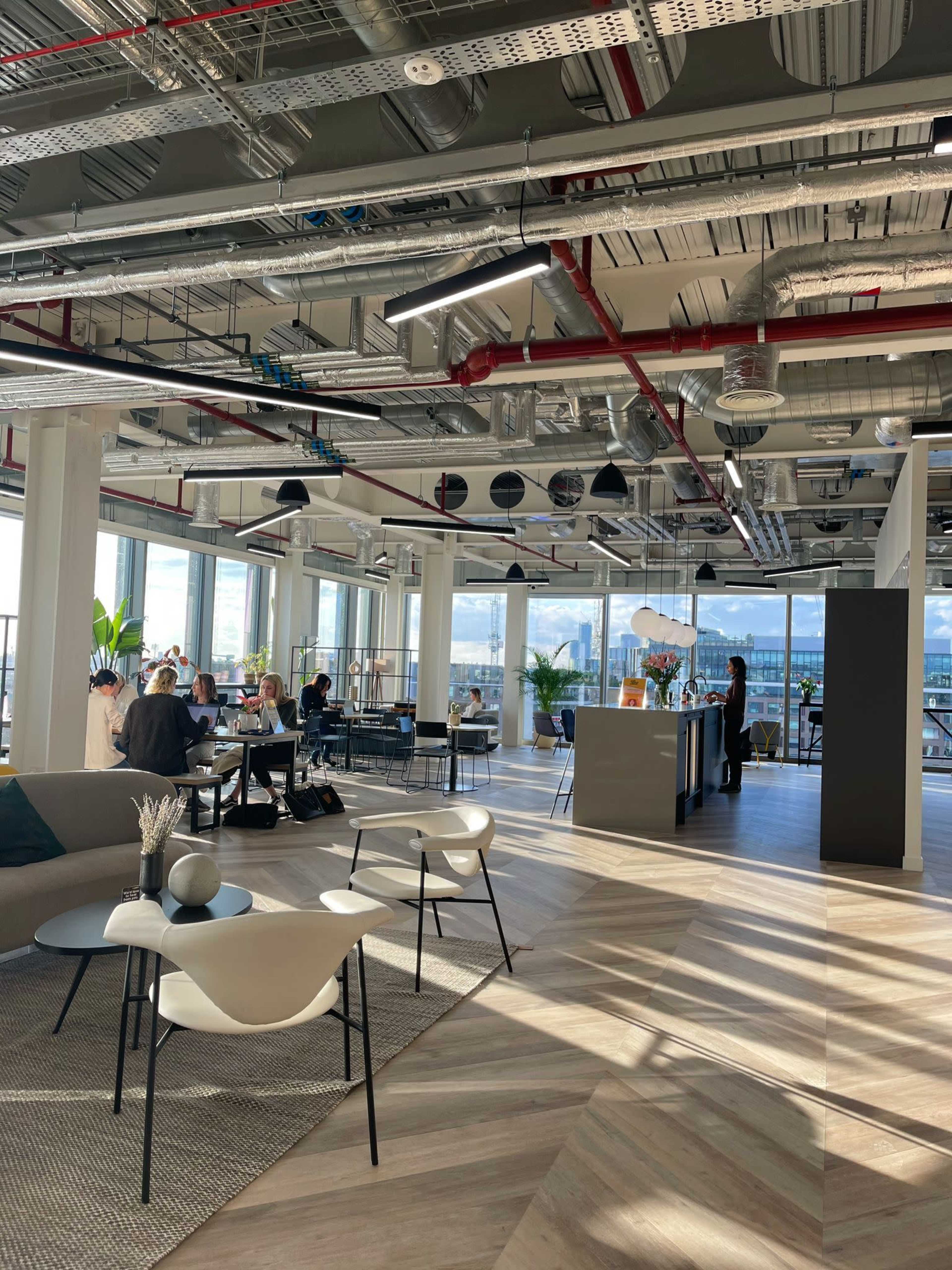 The image shows a modern office space with a seating area, a kitchen counter, and groups of people engaged in conversation near large windows.
