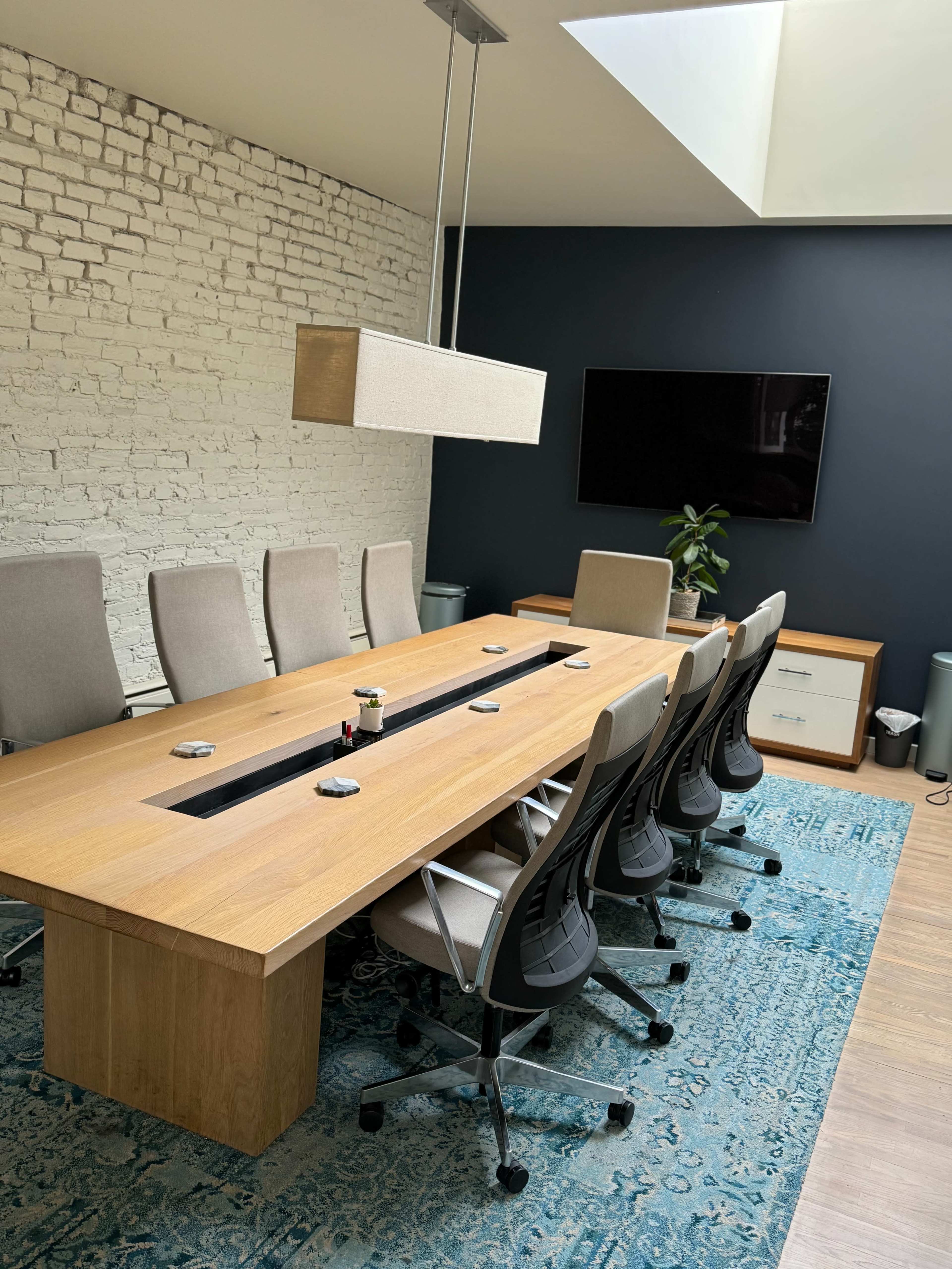 A large wooden conference table is surrounded by ergonomic chairs in a modern meeting room with a skylight and a wall-mounted television.