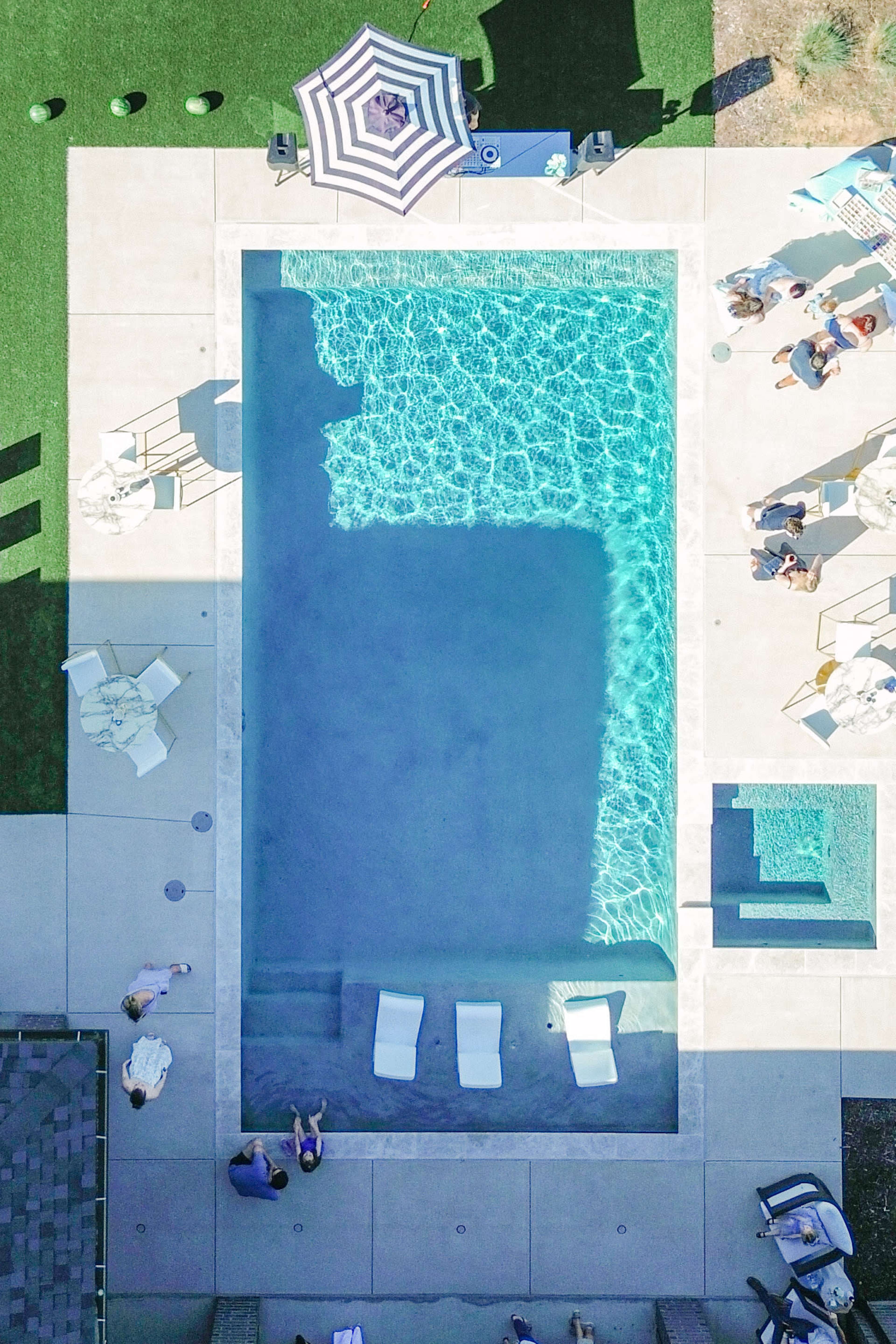 The image shows an aerial view of a rectangular swimming pool surrounded by lounge chairs and an umbrella on a green lawn.