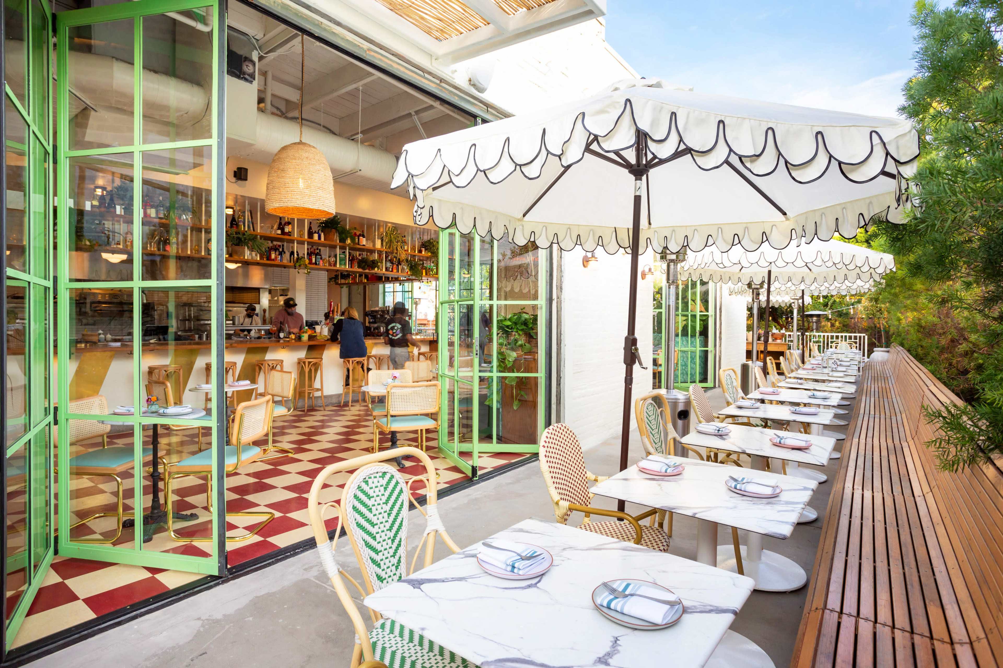The image shows an outdoor dining area of a restaurant with white tables and chairs under large umbrellas, overlooking a bar inside.