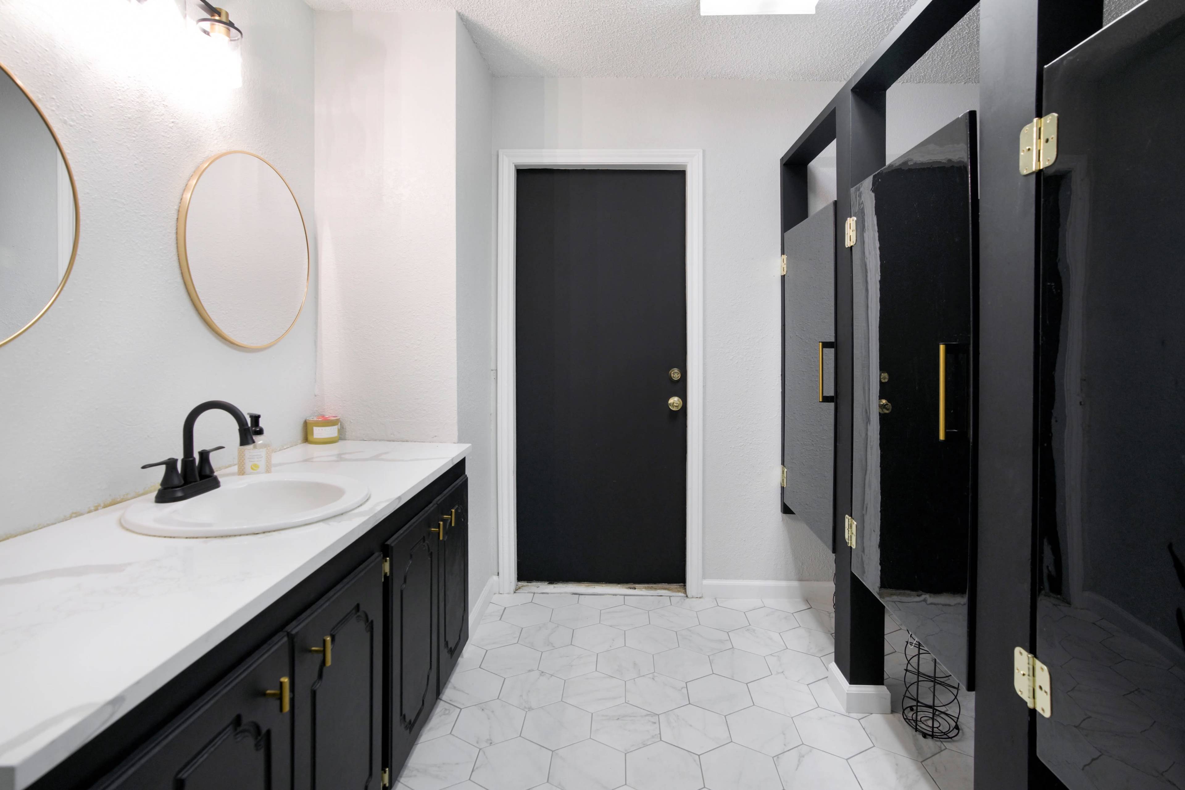 The image shows a modern bathroom featuring a black door, white walls, a circular mirror, a marble countertop sink, and hexagonal floor tiles.