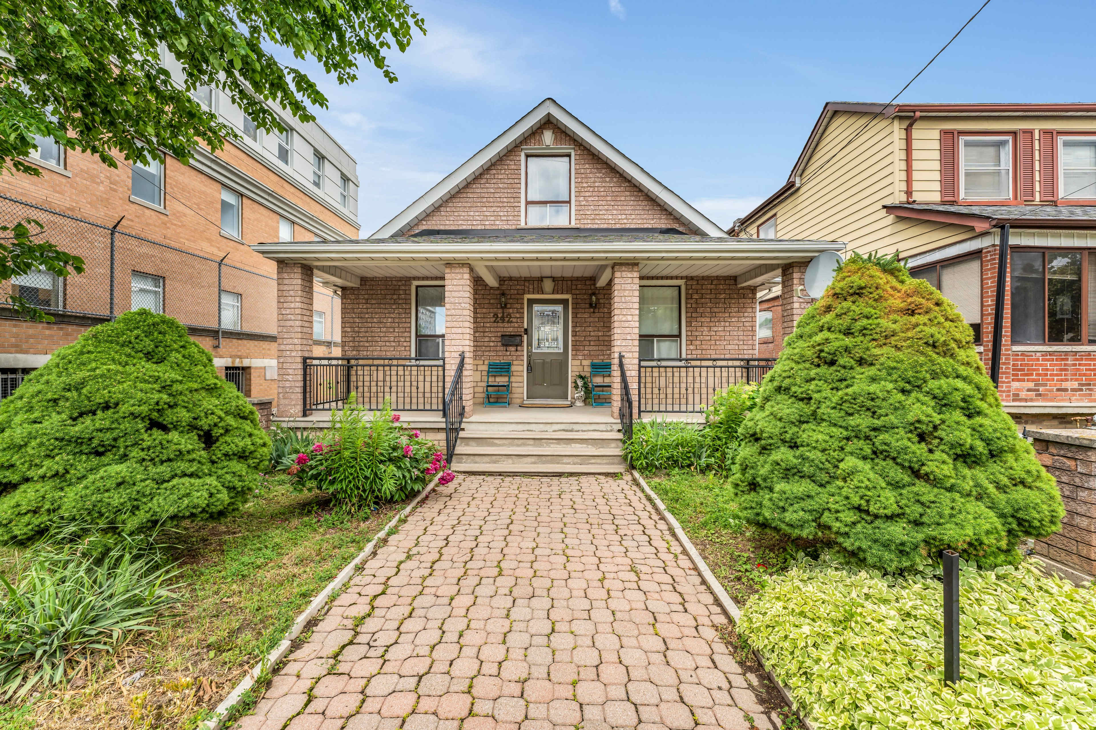 A brick house with a sloped roof is flanked by well-maintained bushes and flowers, set along a paved pathway.