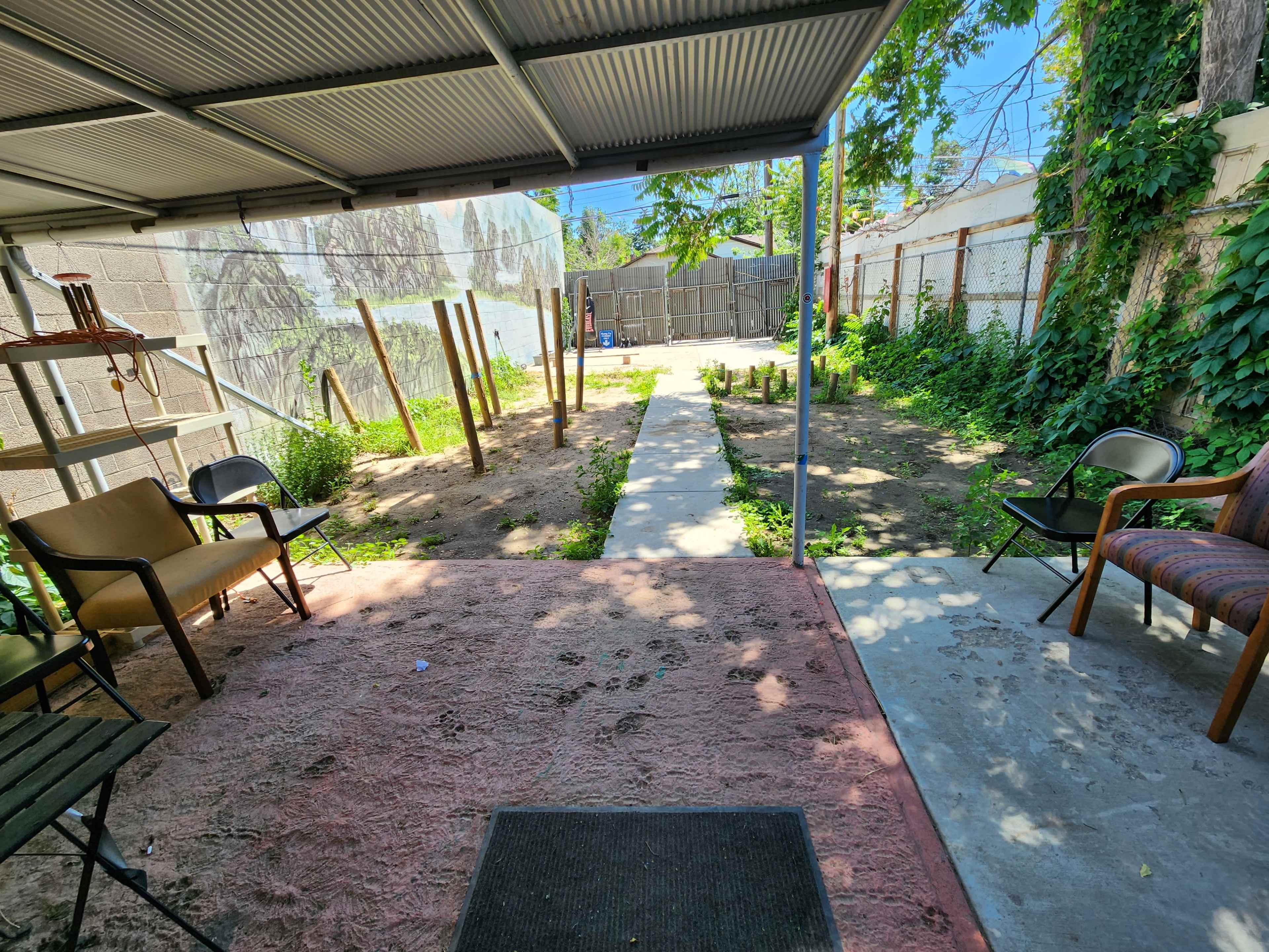 A shaded outdoor area with several empty chairs and a dirt pathway leading to a garden space with wooden poles and greenery in the background.