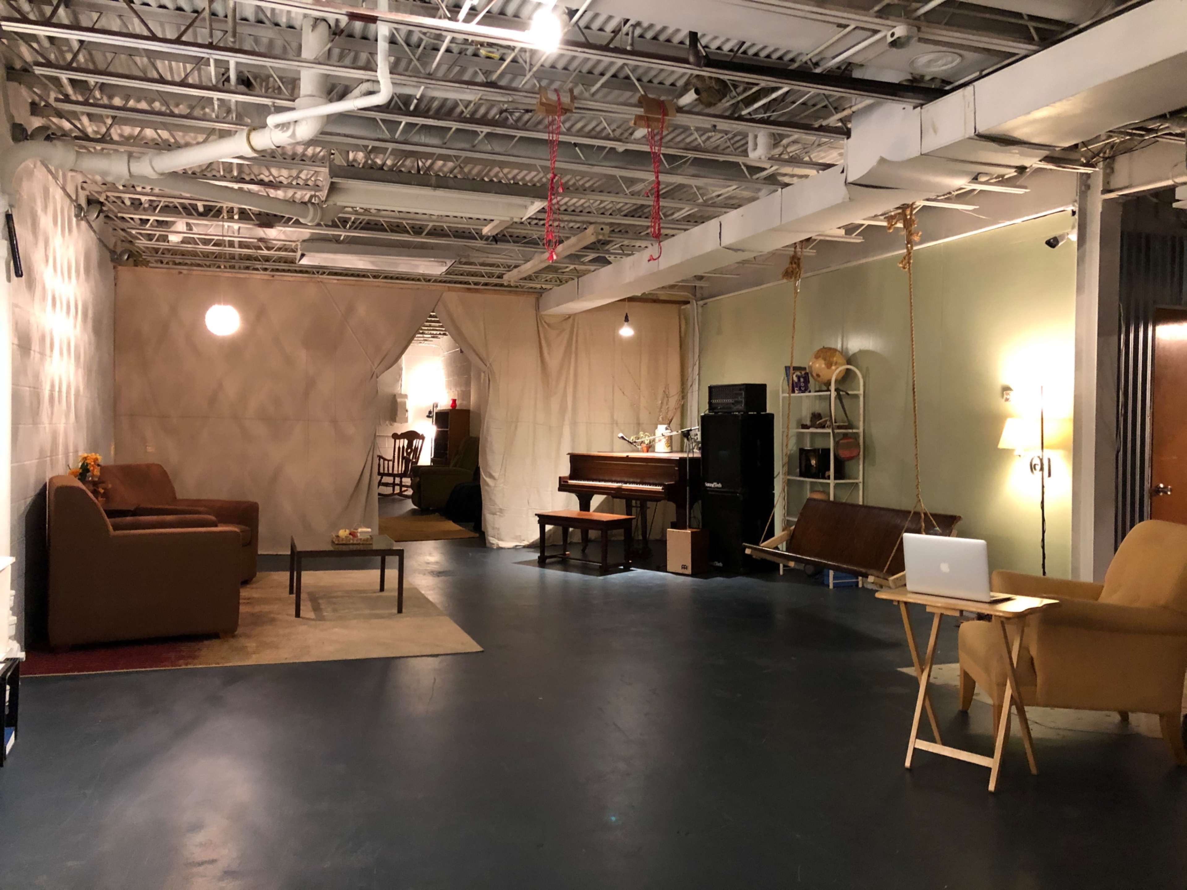 The interior space features a living area with two brown couches, a piano, a swing chair, and a small table with a laptop, all under a ceiling with exposed pipes.