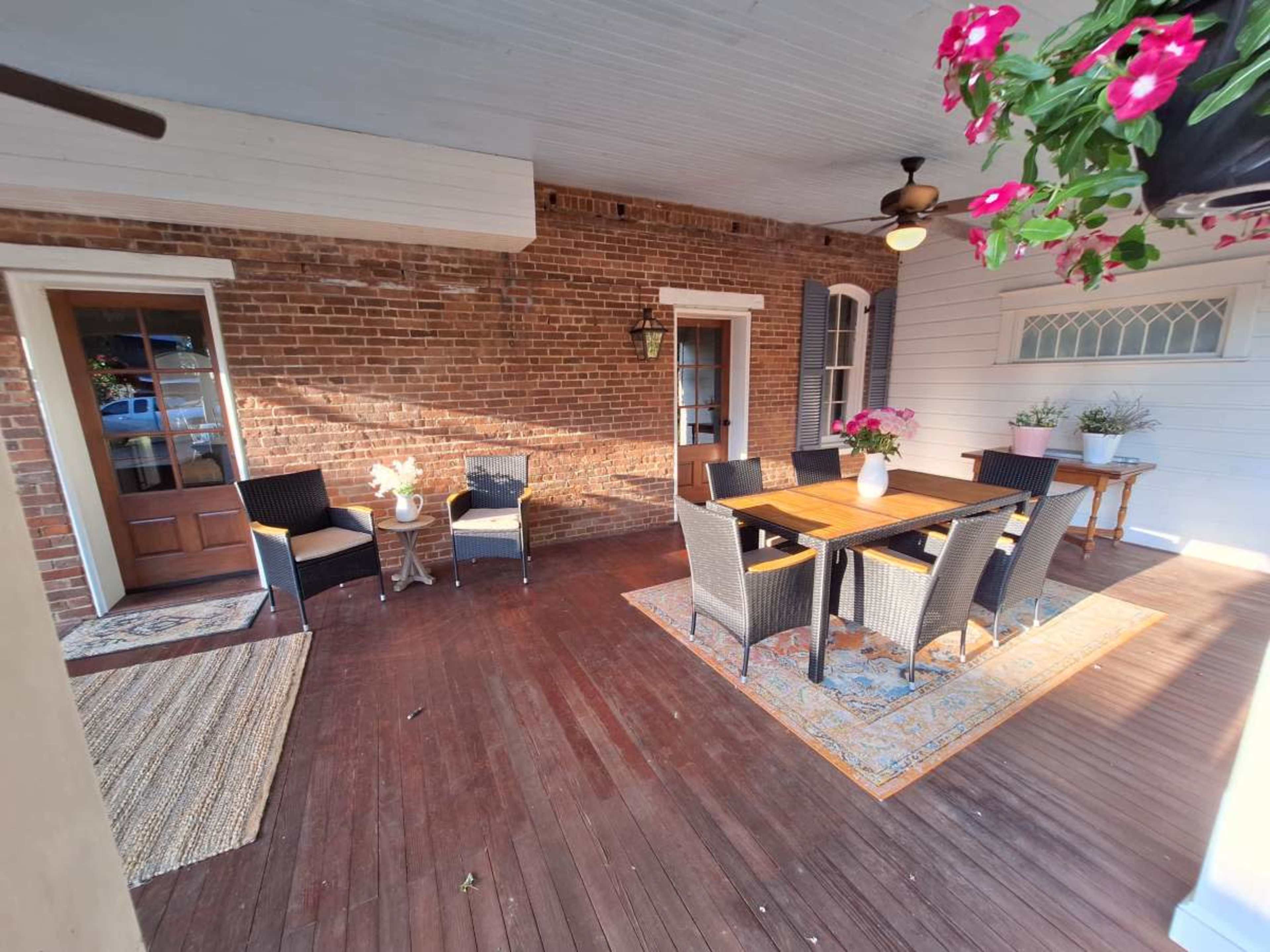 The image shows a covered porch with brick walls, a wooden floor, two seating areas with chairs and a large table surrounded by additional chairs, and potted plants.