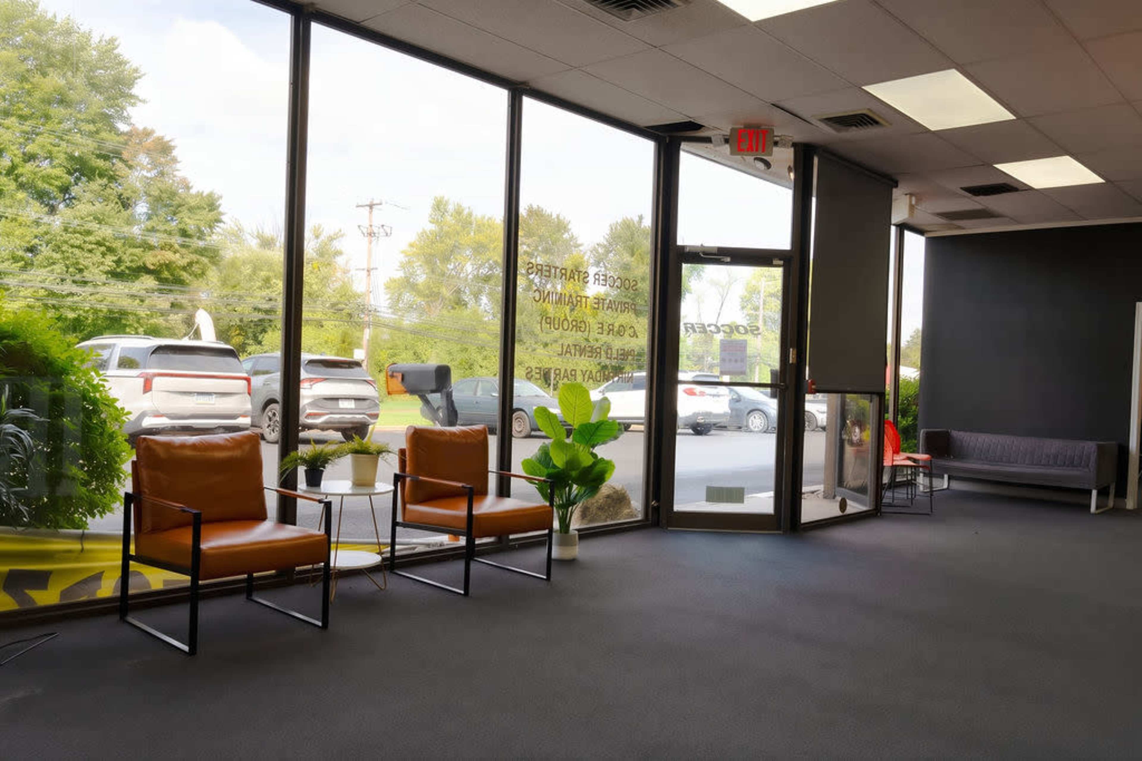 The image shows a modern waiting area with two brown chairs and a potted plant, featuring large windows that allow natural light to illuminate the space.