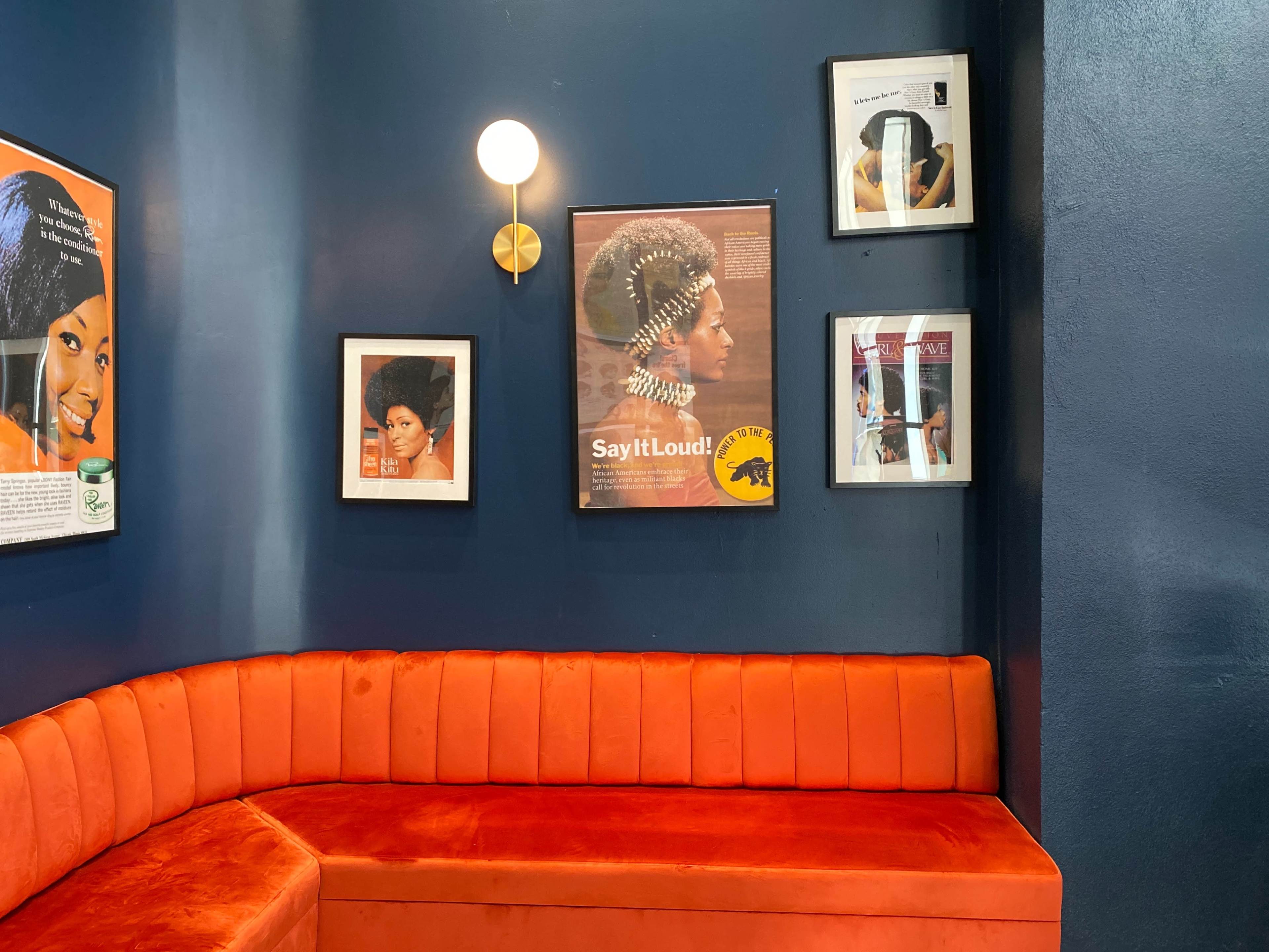 A cozy corner of a café featuring an orange velvet bench against a dark blue wall adorned with framed artwork and photographs.