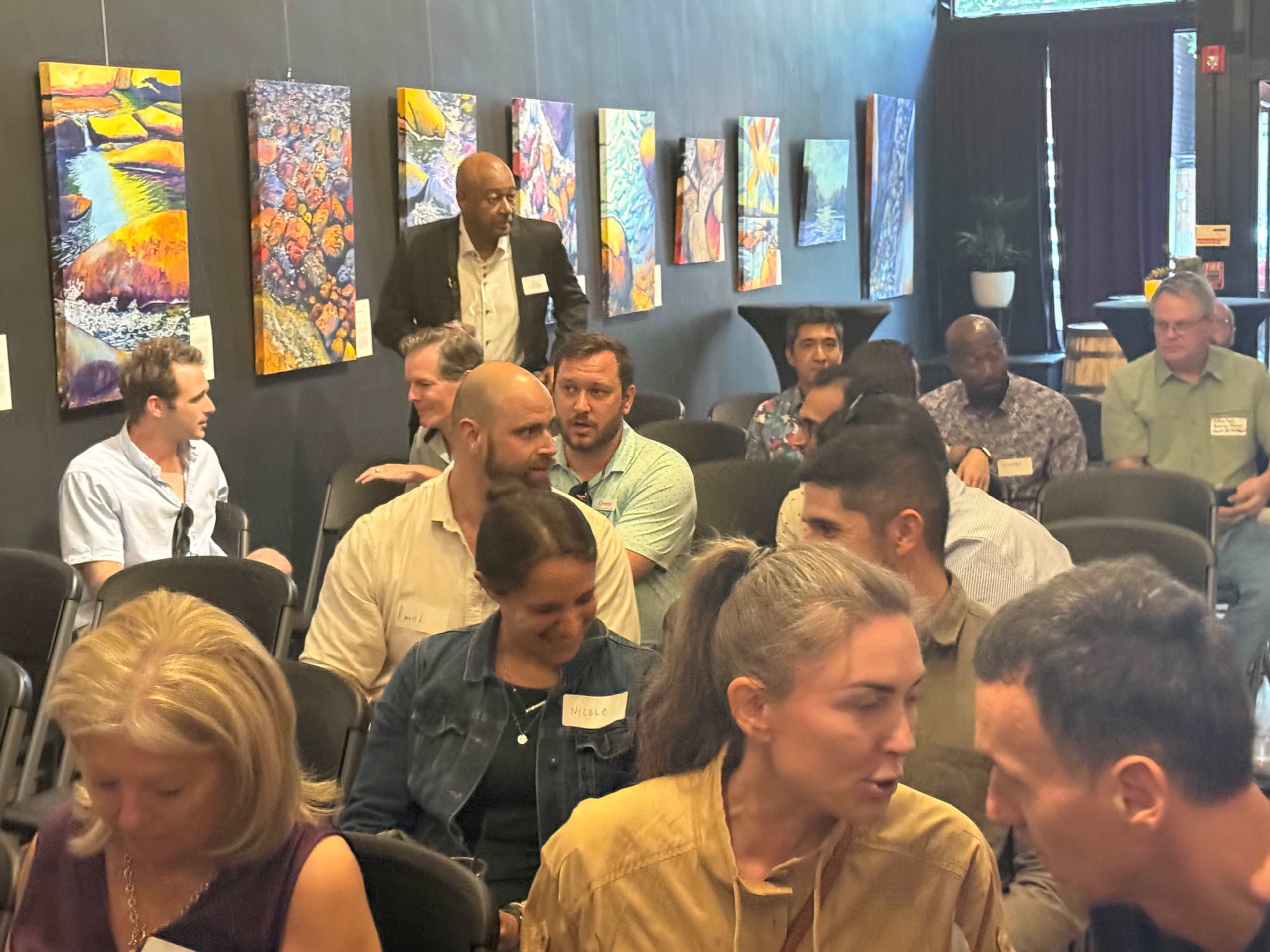 A group of people is seated in a room with colorful artwork displayed on the walls, while one individual stands in the background.