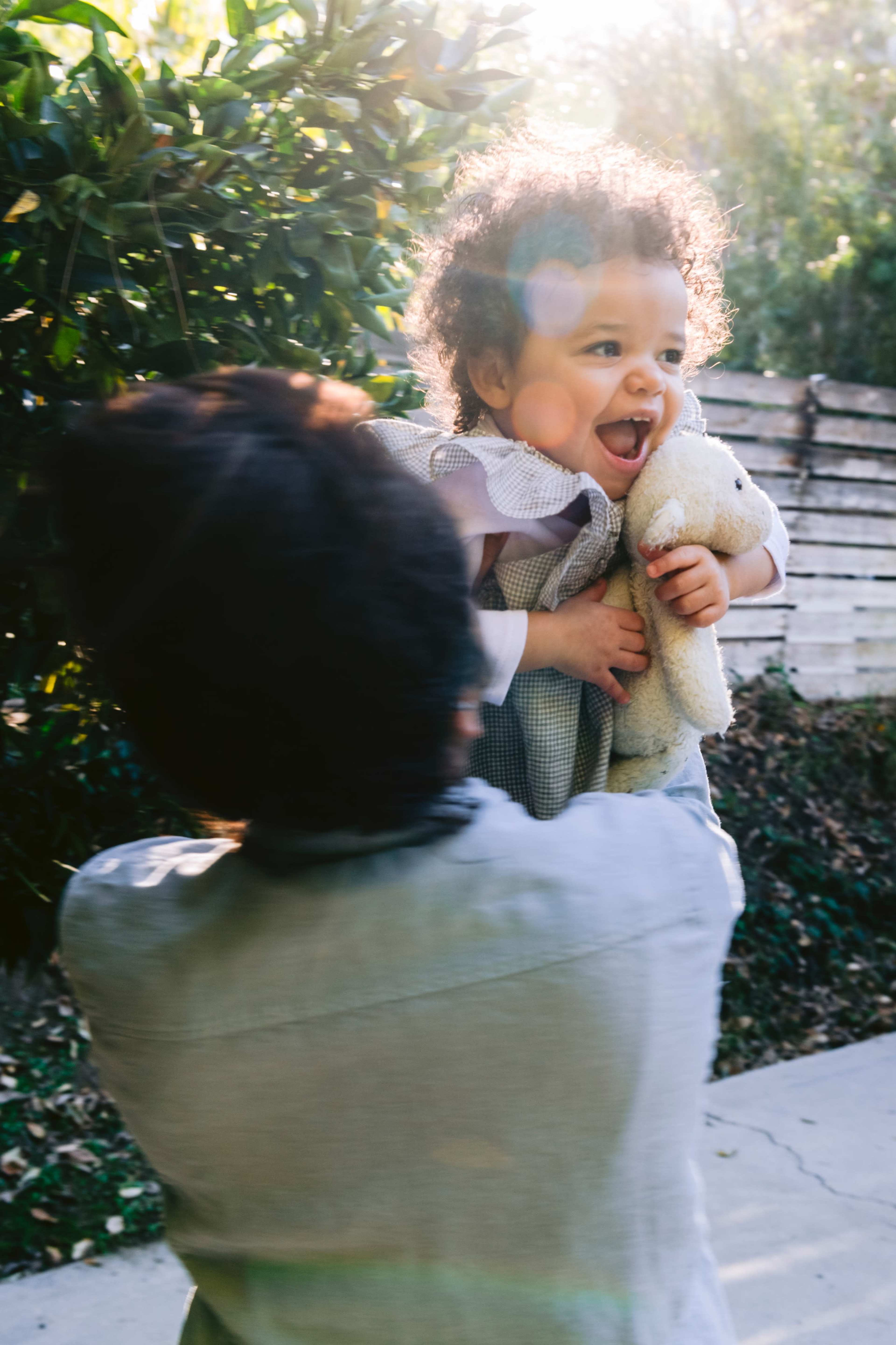 A person holds a smiling child with curly hair and a stuffed animal in a natural outdoor setting.