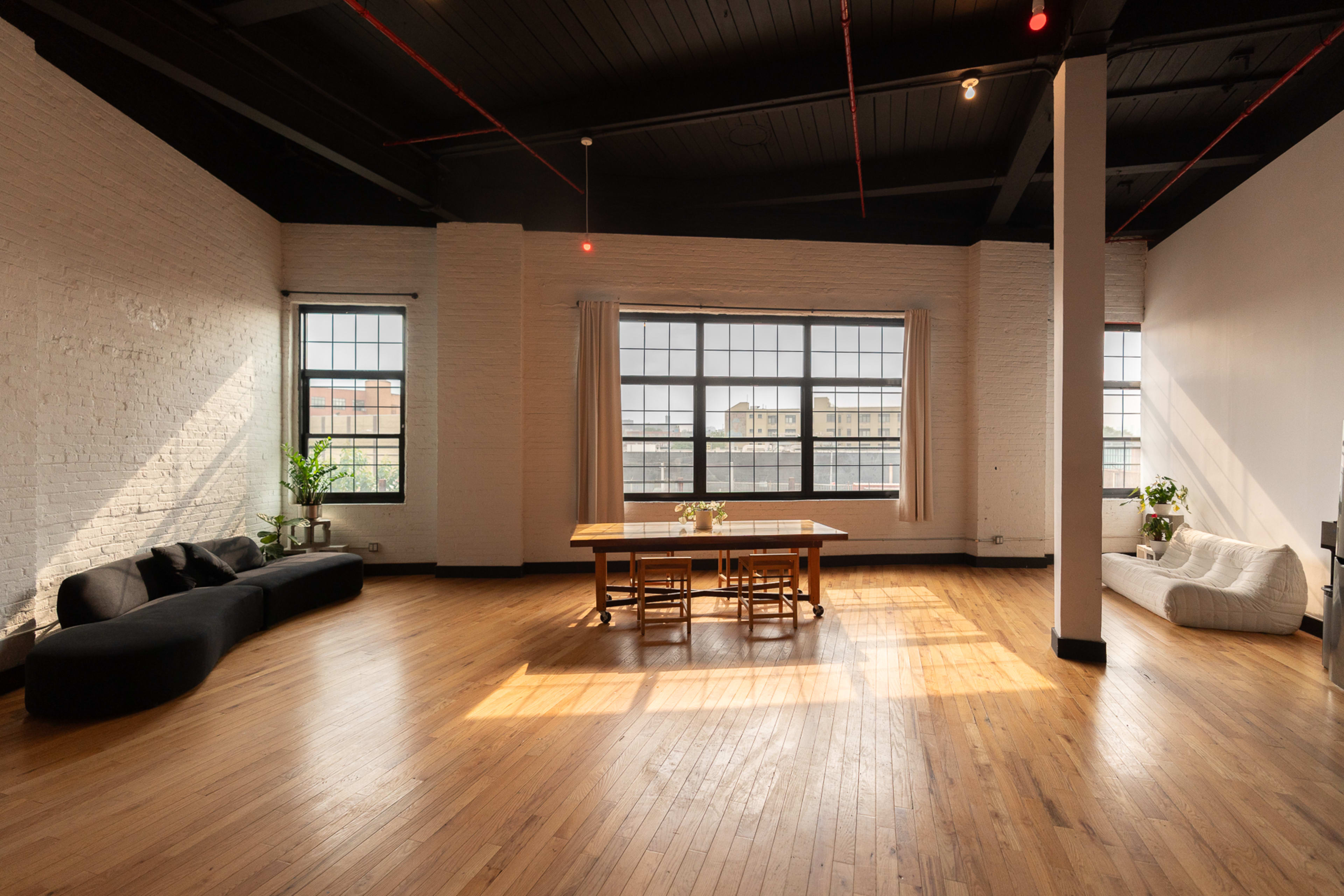 The image shows a spacious, well-lit room with large windows, wooden floors, and a central dining table surrounded by seating arrangements.