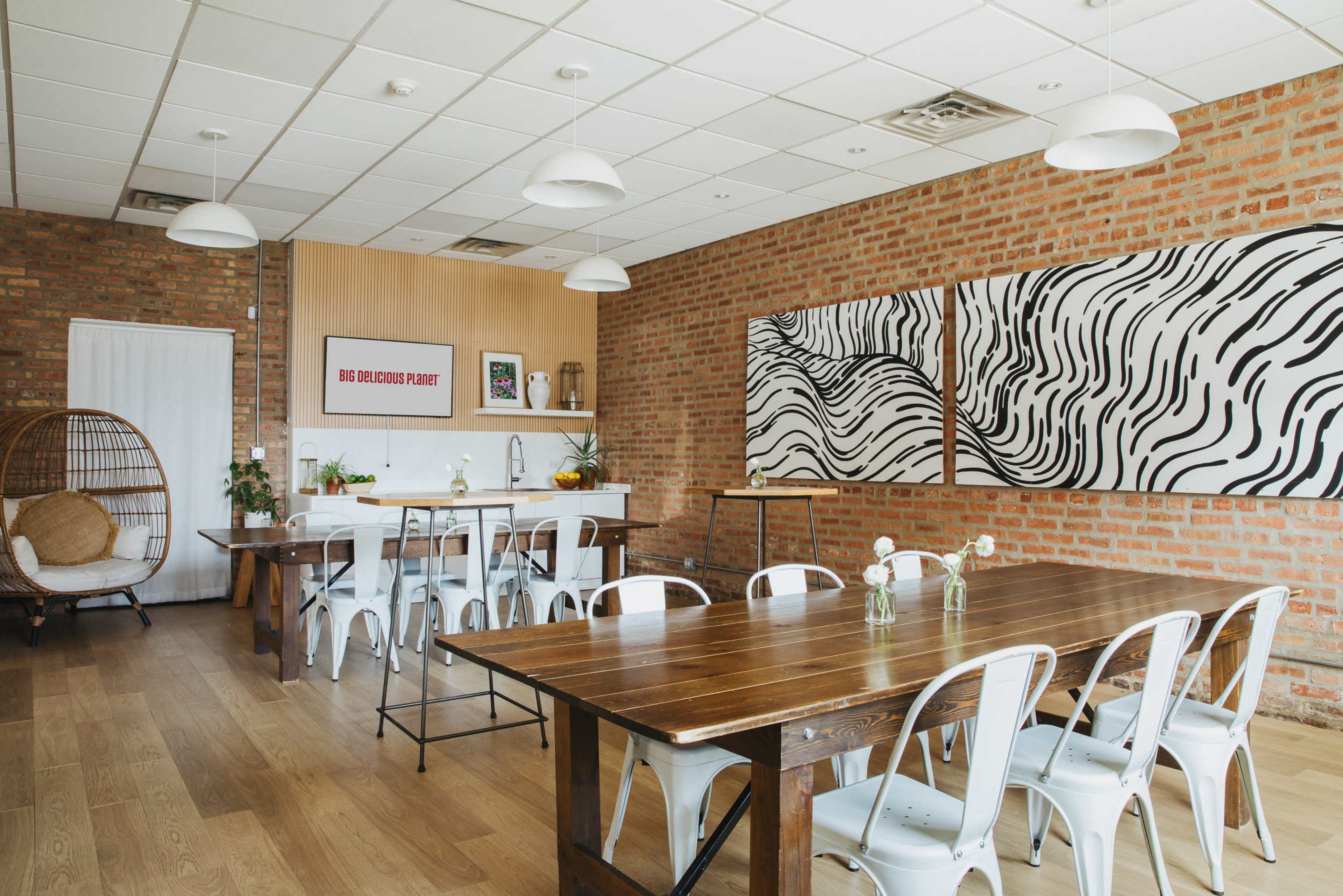 The image shows a modern meeting space with wooden tables, white chairs, a brick wall, and large abstract paintings.