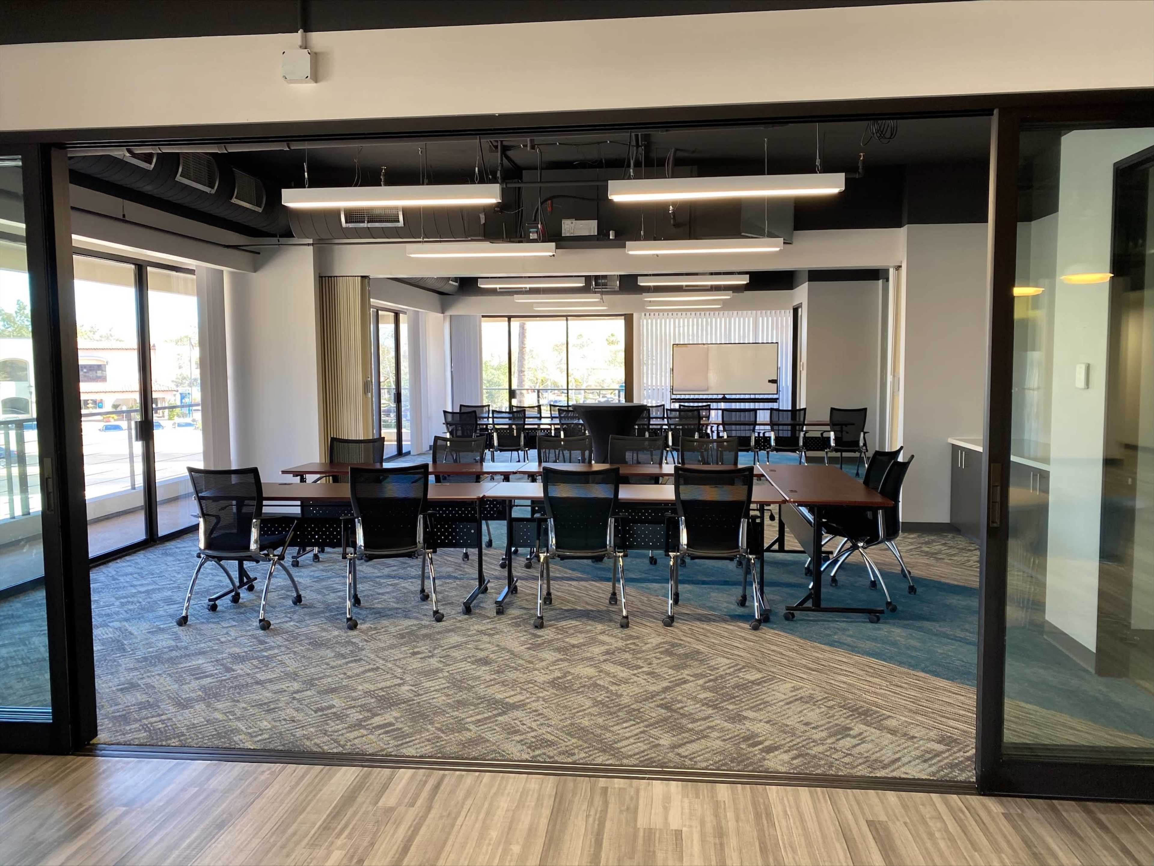 A conference room is set up with multiple tables and chairs, featuring large windows that provide natural light and views of the outdoors.
