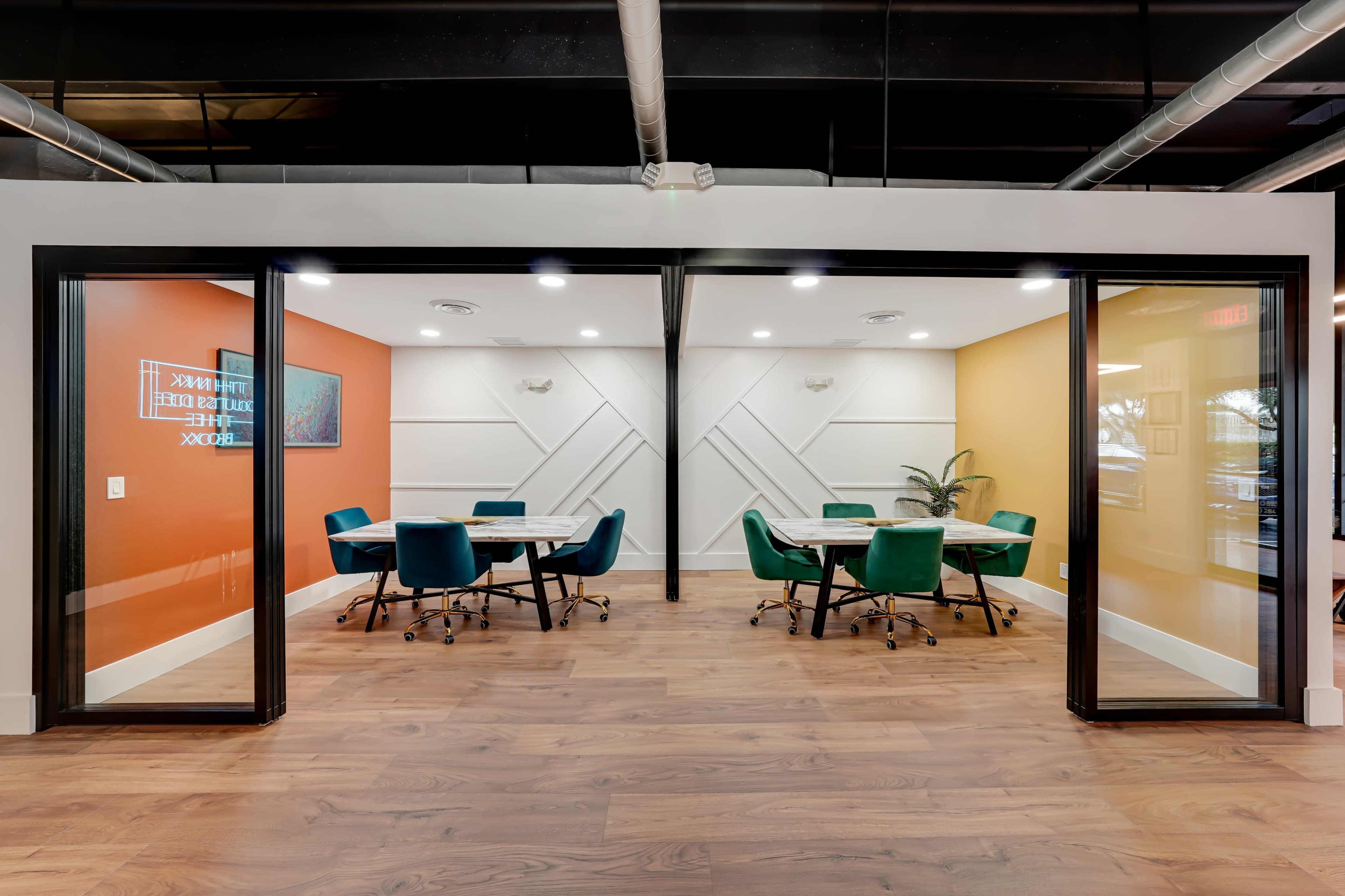 The image shows two meeting rooms with glass walls, each containing a round table surrounded by teal chairs, set against orange and yellow accent walls.