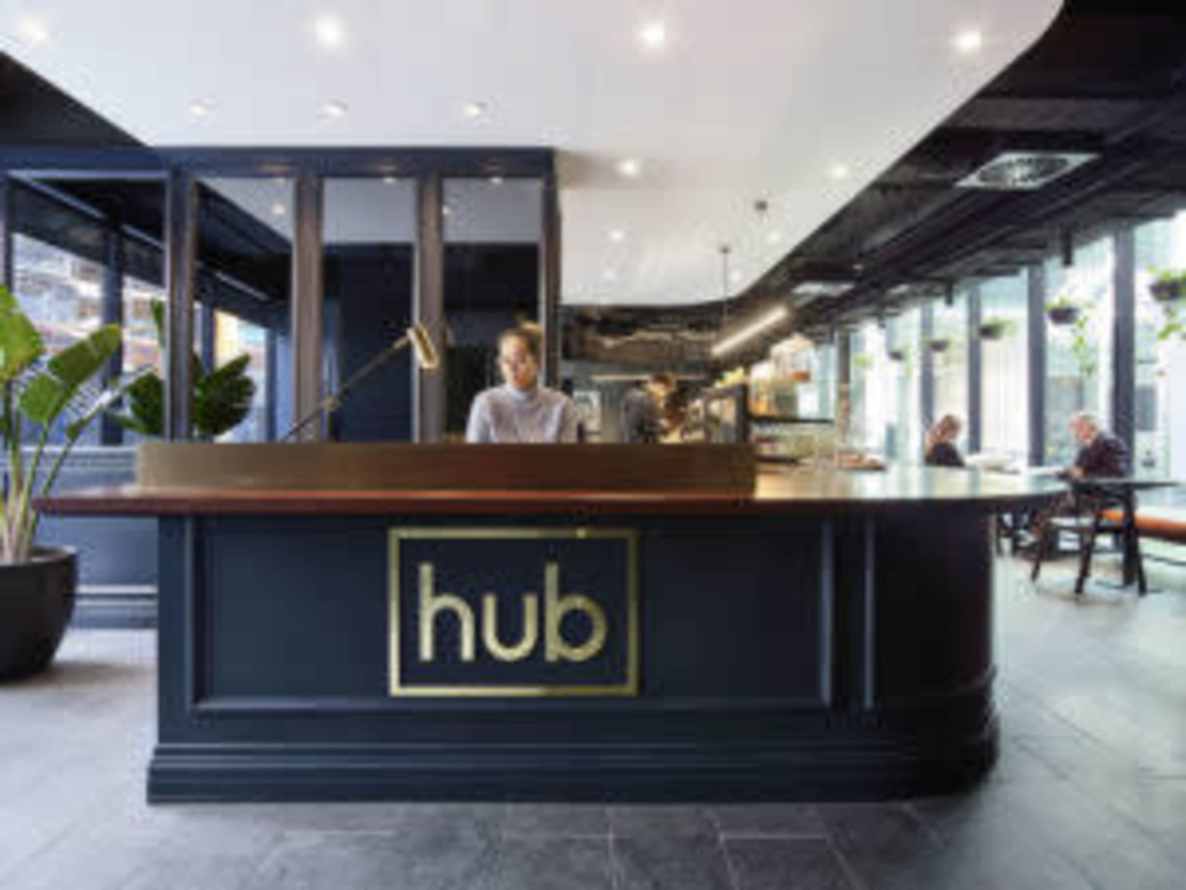 A sleek reception desk with the word "hub" prominently displayed, staffed by a person, in a modern, well-lit interior with seating areas in the background.
