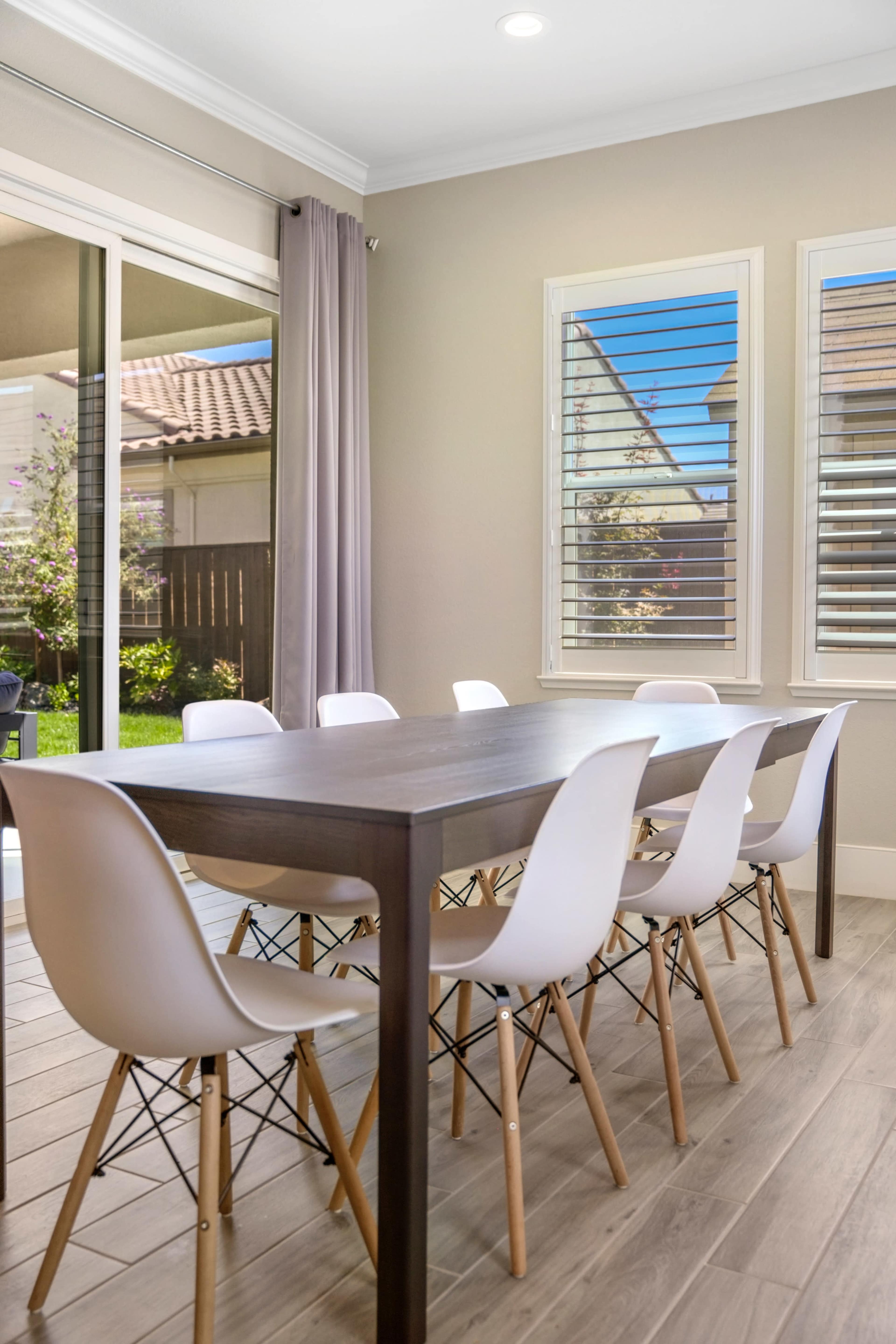 A modern dining area features a long wooden table surrounded by white chairs, with large windows showing a view of an outdoor space.