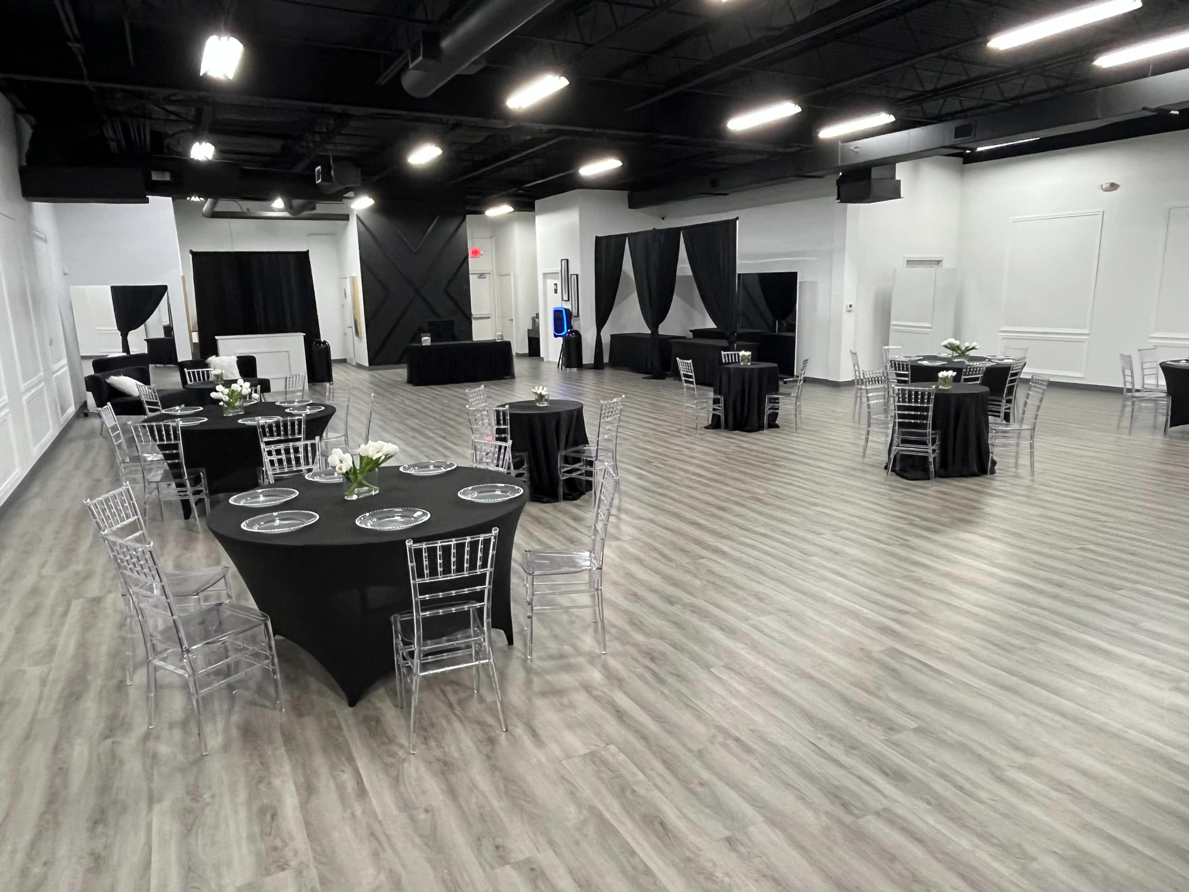 The image shows a spacious event venue with several round tables dressed in black tablecloths and clear chiavari chairs, set up for a gathering.