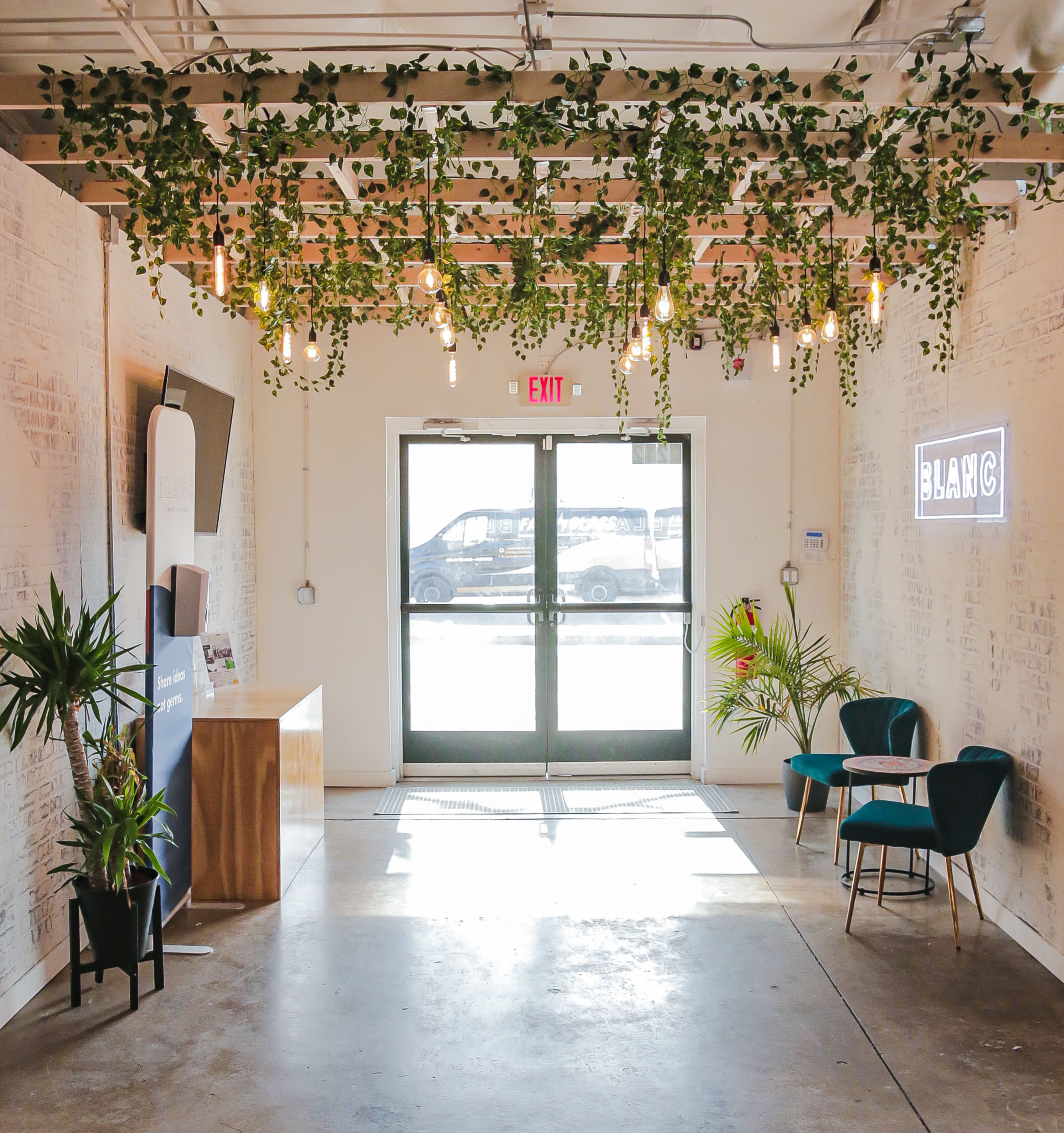 The image shows a modern entryway with a glass door, green plants hanging from the ceiling, and two blue chairs by the wall.