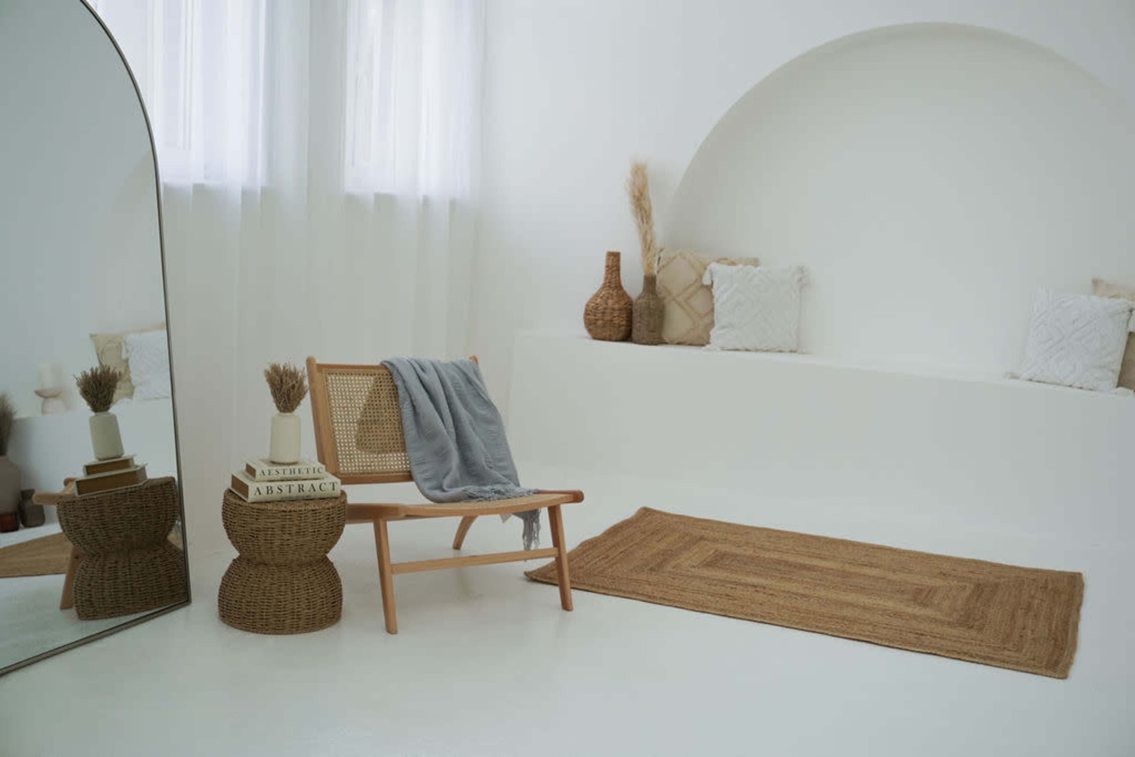 The image depicts a minimalist room featuring a chair, a woven rug, decorative pillows, and large windows allowing natural light to enter.