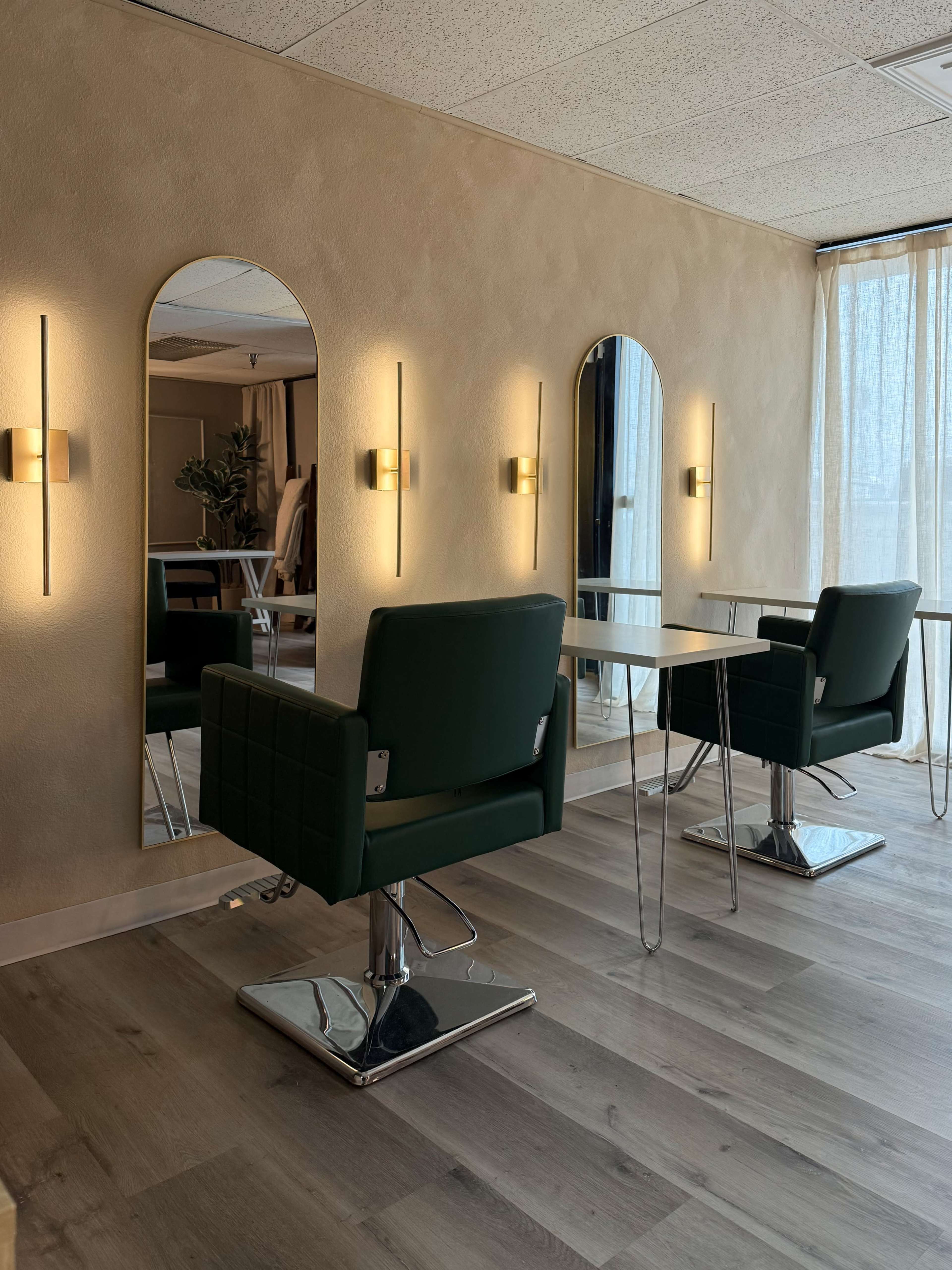 The image shows a modern hair salon with two green styling chairs, three framed mirrors, and warm wall lights, all set in a well-lit room with wooden flooring.