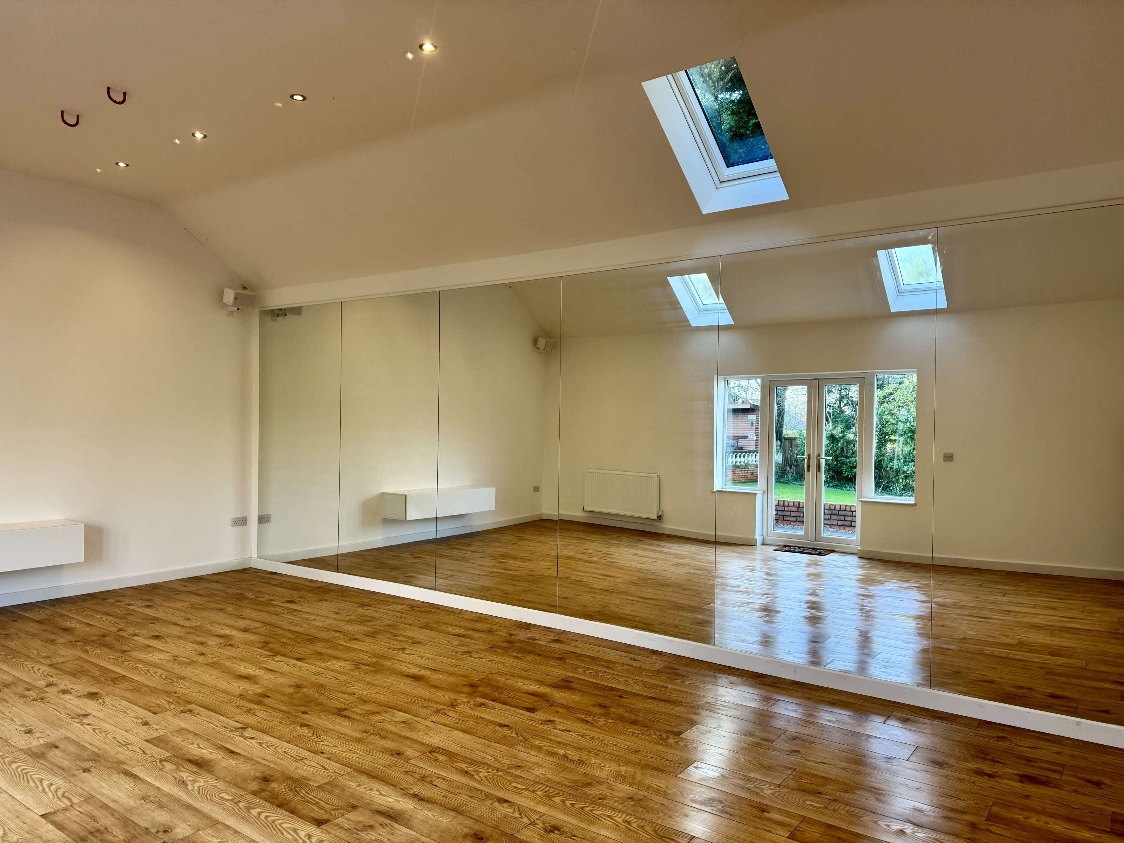A spacious room with wooden flooring and a large wall of mirrors, featuring skylights and a door that opens to an outdoor area.